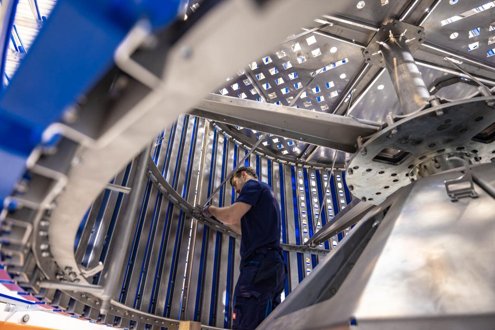 A worker in a blue uniform inspects or assembles a large, circular industrial machine with metal beams and panels in a factory setting.
