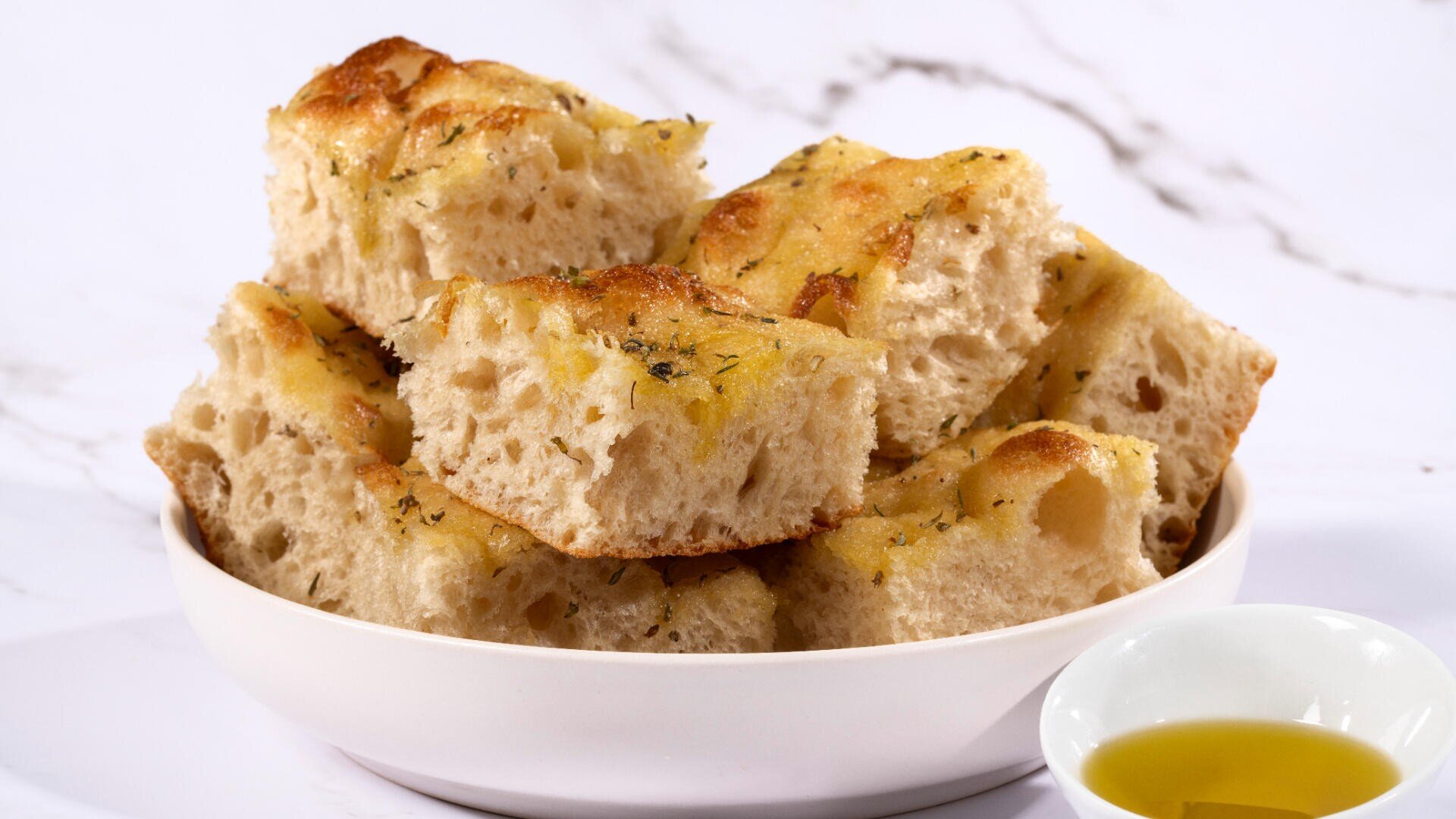 A white bowl filled with thick, golden-brown slices of focaccia bread seasoned with herbs, next to a small dish of olive oil, against a white marble background.