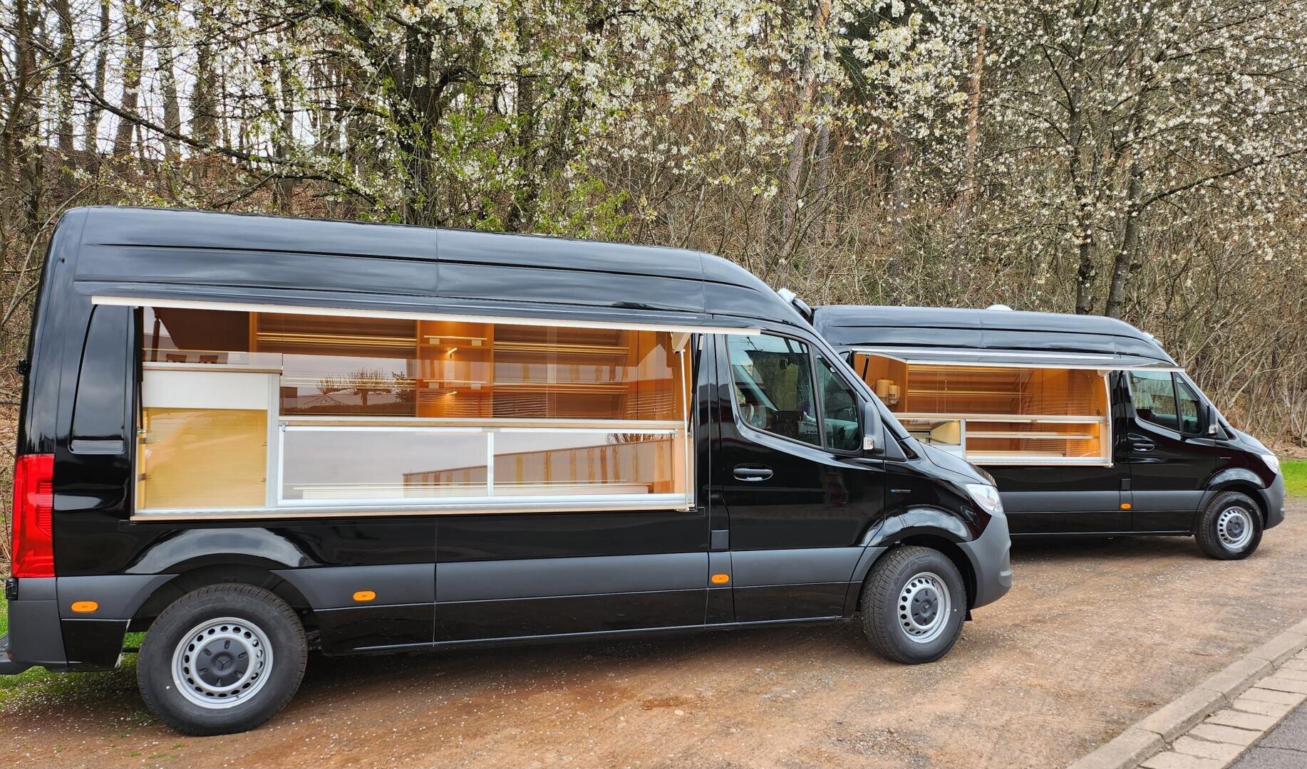Zwei schwarze Lieferwagen mit großen Seitenfenstern, die den Blick auf ein modernes, gut beleuchtetes Holzinterieur freigeben, sind auf einem Feldweg neben blühenden Bäumen geparkt. Die Innenräume scheinen für mobile Geschäfts- oder Vorzeigezwecke konzipiert zu sein.