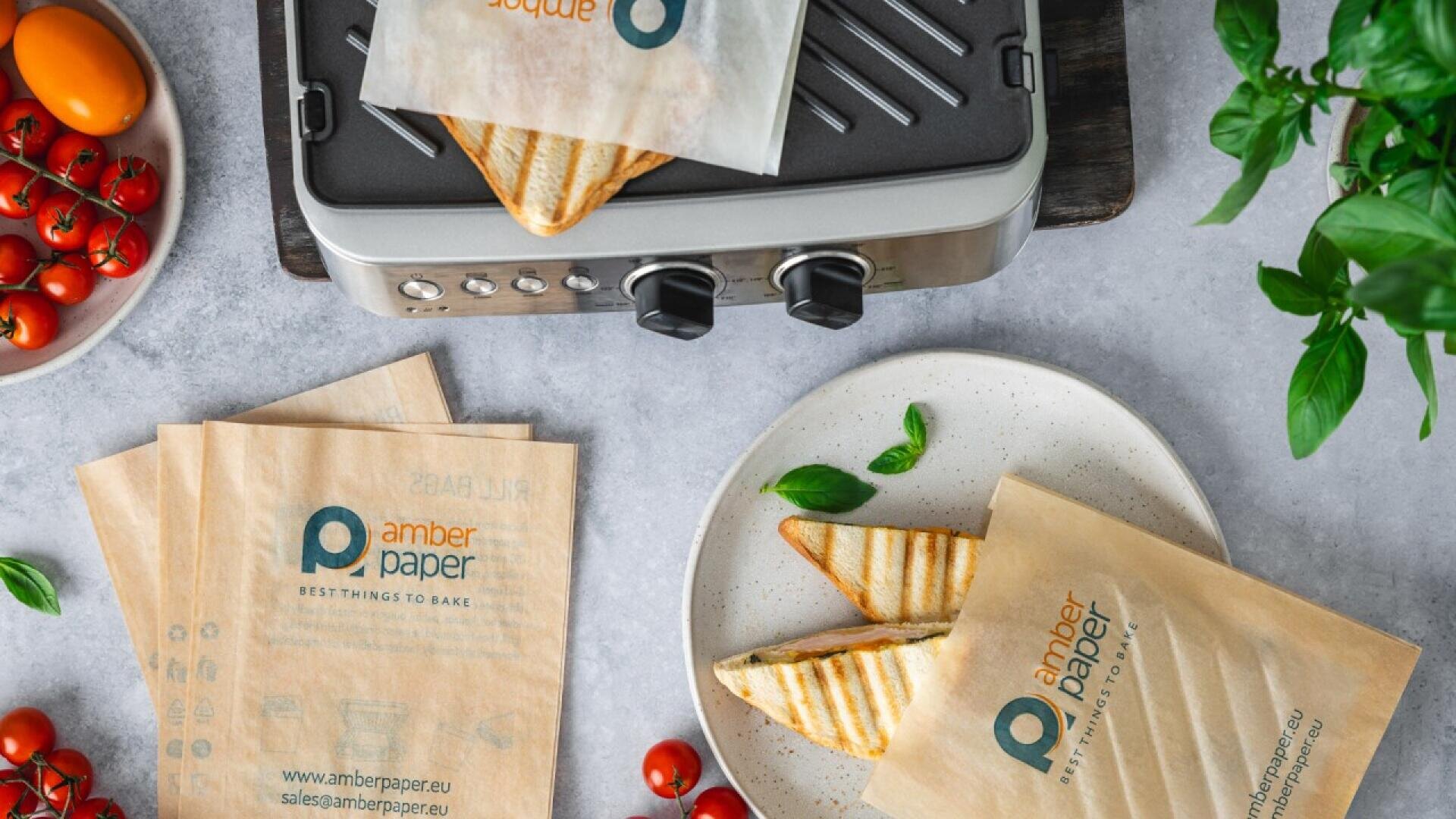 Zwei getoastete Sandwiches in braunem Pergamentpapier liegen auf einem weißen Teller neben einem Grill. Frisches Basilikum, Tomaten und weitere Pergamentblätter umgeben den Teller auf einer hellgrauen Fläche.