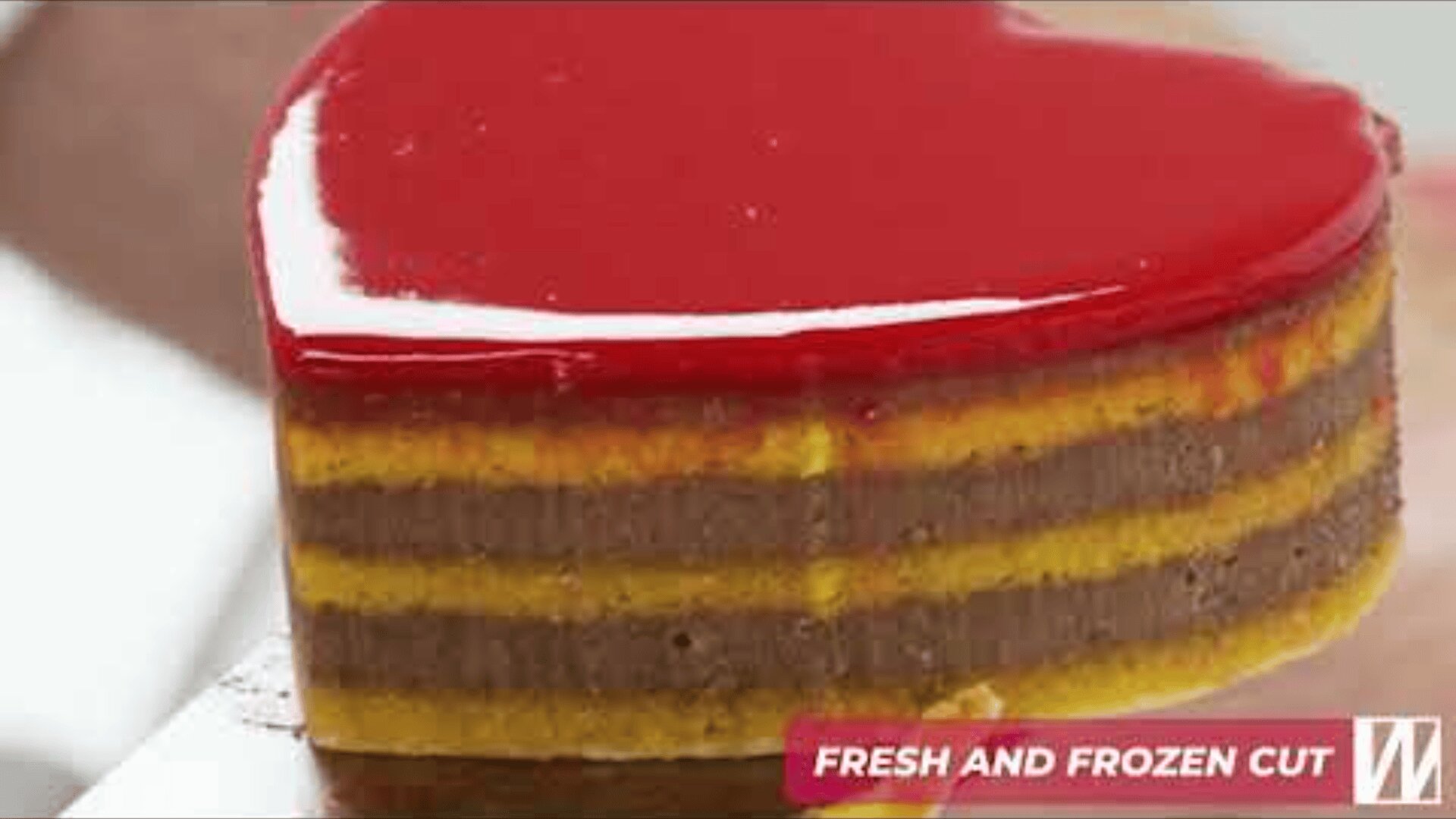 Ein herzförmiges Schichtdessert mit glänzender roter Glasur und abwechselnd gelben und braunen Schichten, gehalten auf einem Spatel. In der Ecke steht „FRESH AND FROZEN CUT“.