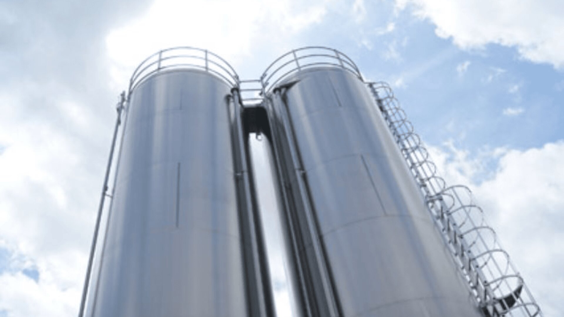 Zwei hohe, zylindrische Metallsilos mit Leitern und Geländern sind nebeneinander aufgestellt und spiegeln den wolkenverhangenen Himmel über ihnen wider.