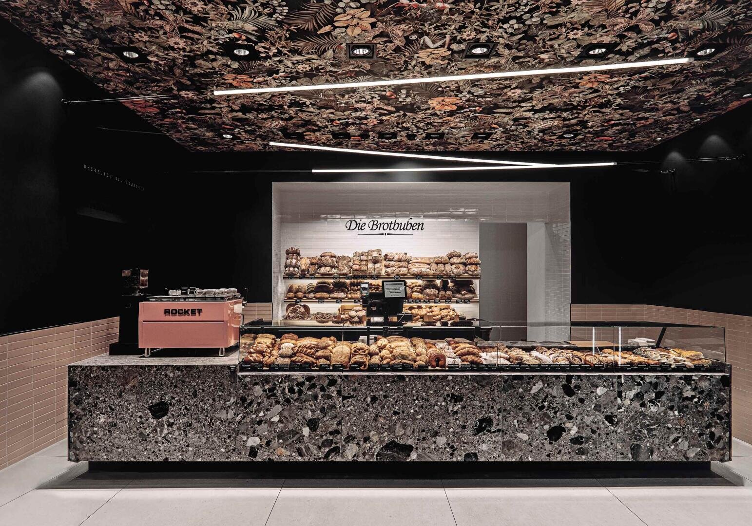 A modern bakery with a terrazzo counter displaying assorted pastries, a pink espresso machine, and shelves filled with bread. The ceiling has a dark floral pattern and the sign above the bread reads Die Brotbuben.