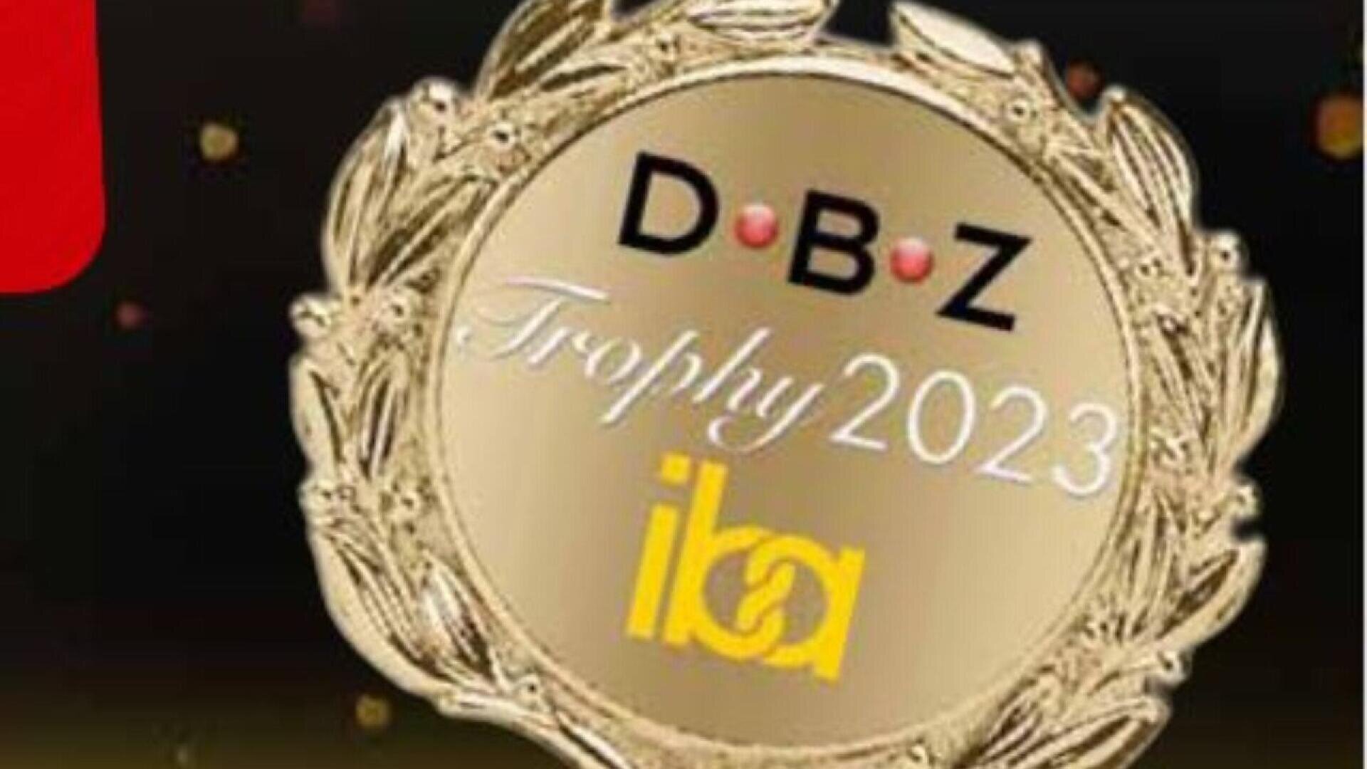 A gold trophy medallion with the text D B Z Trophy 2023 iba embossed in the center, surrounded by a laurel wreath design on a dark background with light spots.