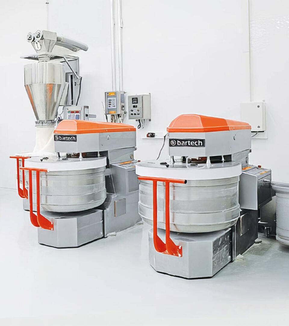 Two large industrial dough mixers with orange lids and stainless steel bowls stand in a clean, white room. Control panels and a stainless steel hopper can be seen in the background.