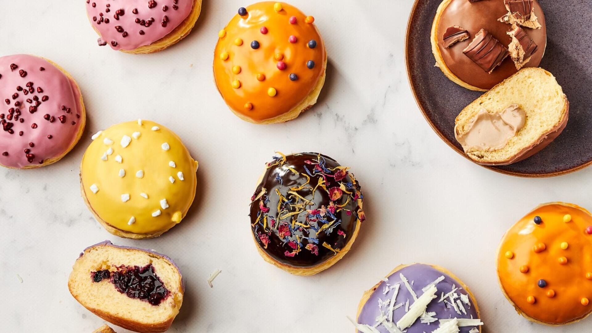 Verschiedene bunte, mit Creme gefüllte Donuts, von denen einige in Scheiben geschnitten sind, um den Blick auf die verschiedenen Füllungen freizugeben, sind auf einer weißen Marmoroberfläche angeordnet. Auf der rechten Seite steht ein Teller mit zwei Donuts.