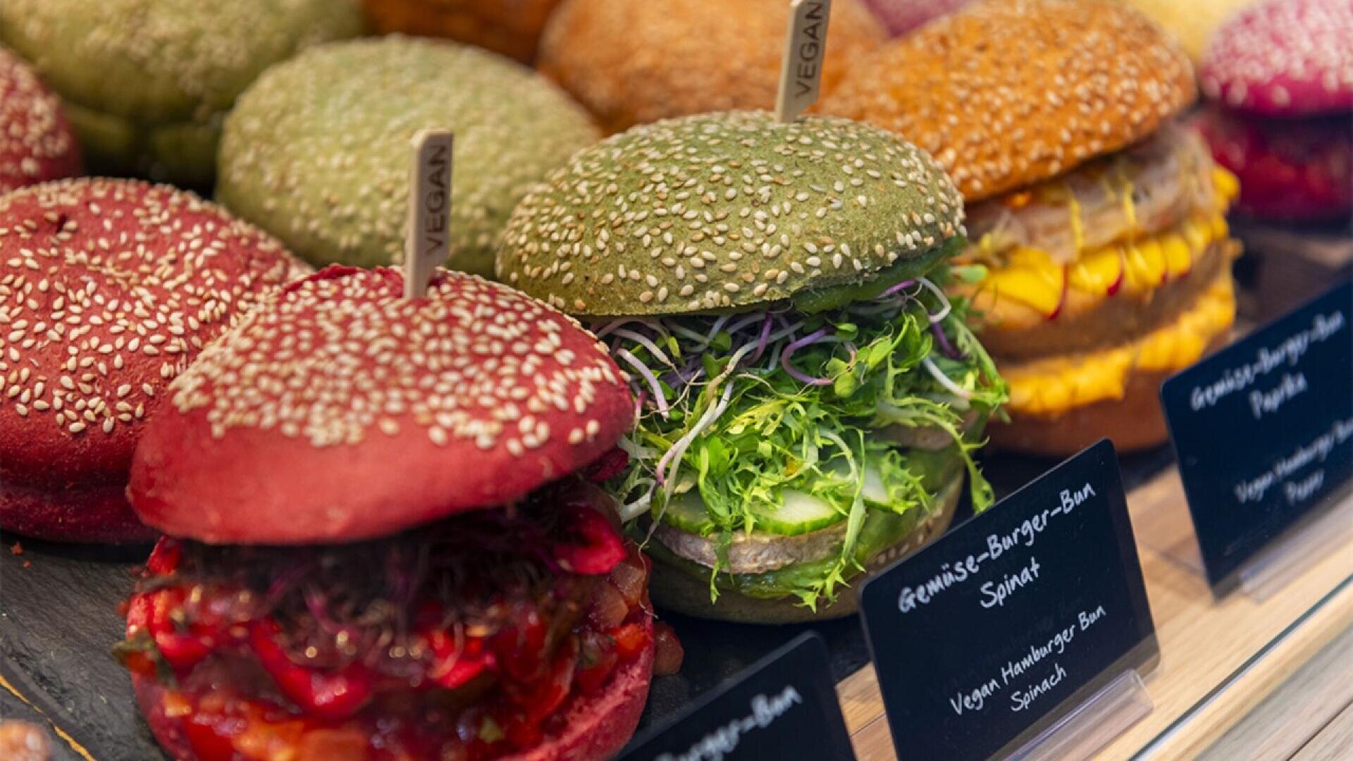 A display of colorful vegan burgers with red, green, and yellow sesame seed buns, filled with various fresh vegetables and greens. Small signs and labels indicate the burgers are vegan.
