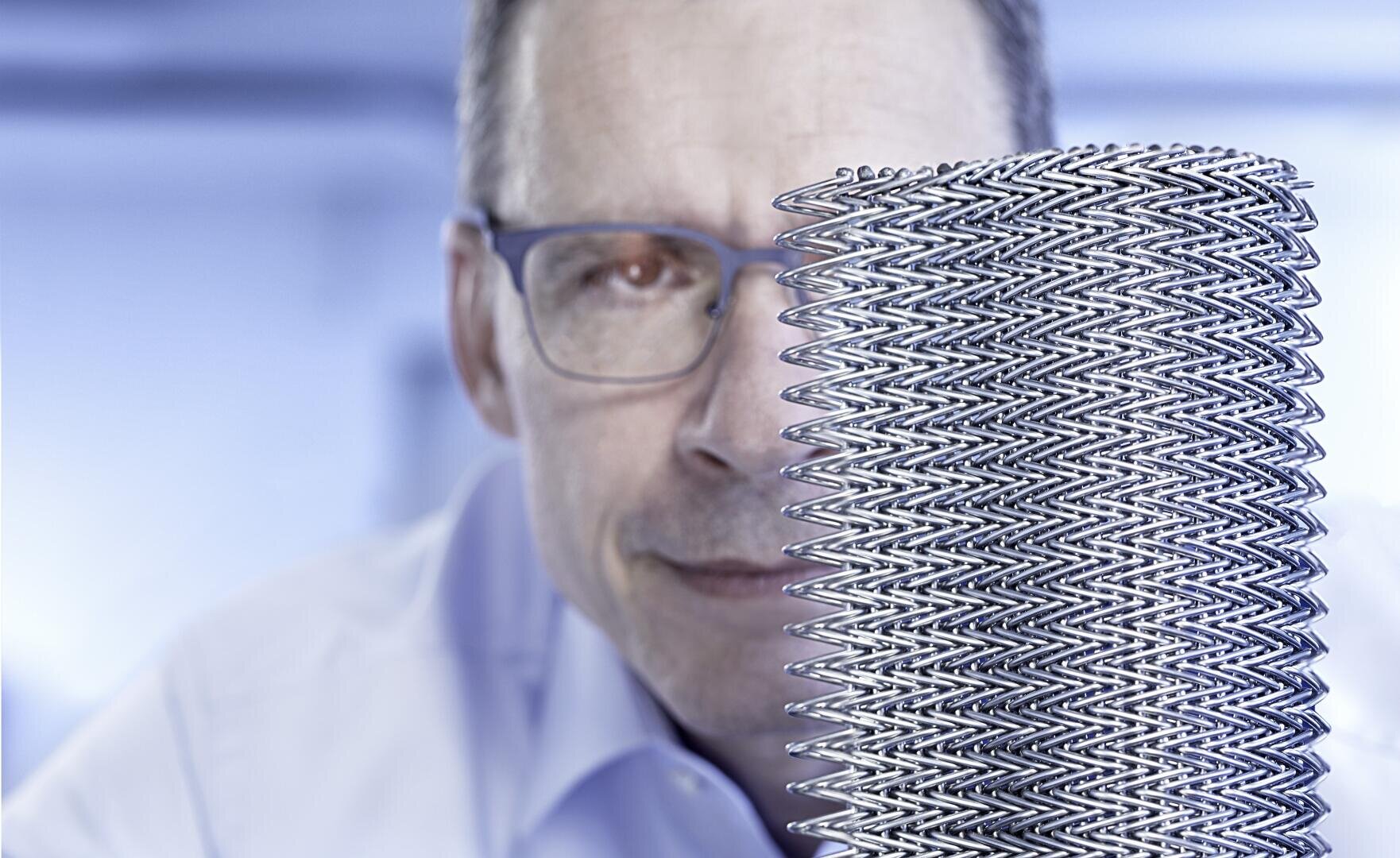 Ein Mann mit Brille blickt auf ein großes, zylindrisches Metallgeflecht, das im Vordergrund zu sehen ist, mit einem blau getönten, verschwommenen Hintergrund.
