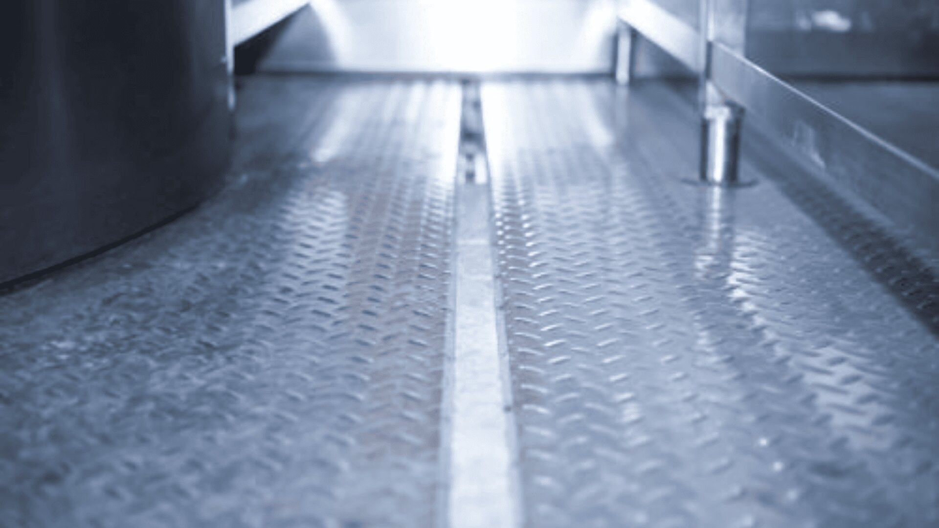Close-up view of a metal diamond plate floor with a narrow central groove, reflecting cool light from above, and surrounded by metallic structures on both sides.