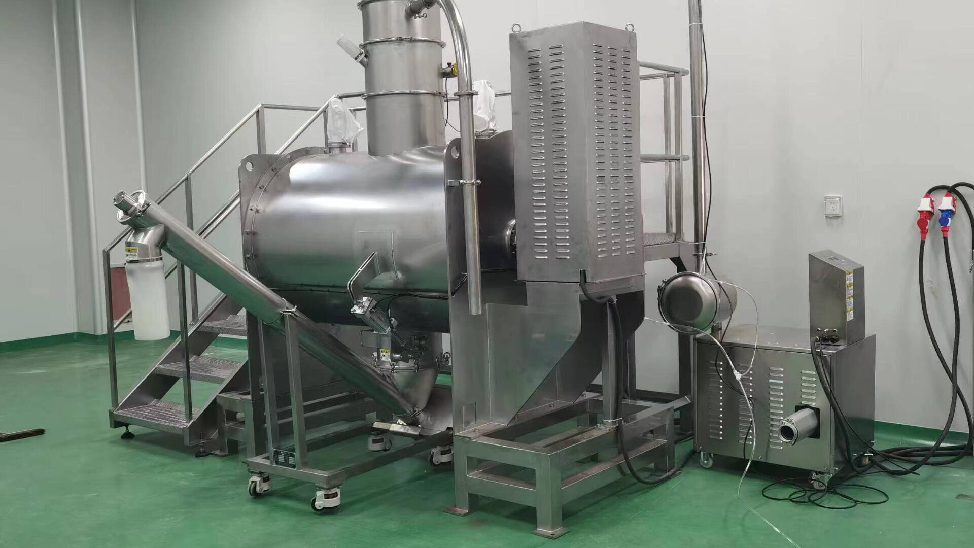 Large stainless steel industrial machine with cylindrical chamber, control panel, pipes, and stairs, set in a clean room with green flooring and white walls. Equipment for processing or manufacturing.