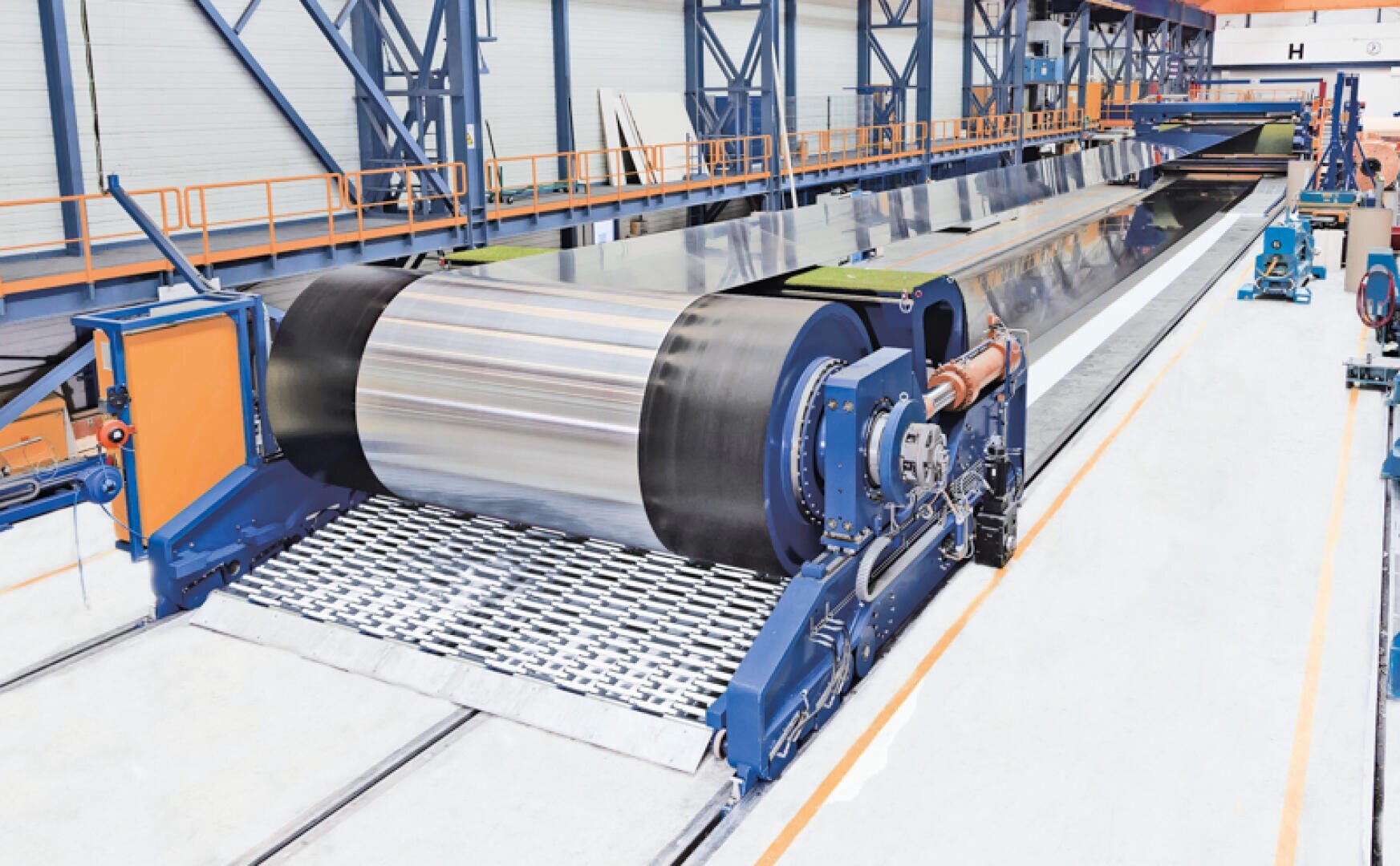 Eine große Industriemaschine in einer Fabrik rollt ein Blech aus glänzendem Metall auf ein Fördersystem, umgeben von blauen und orangefarbenen Metallgeländern und Stützen.