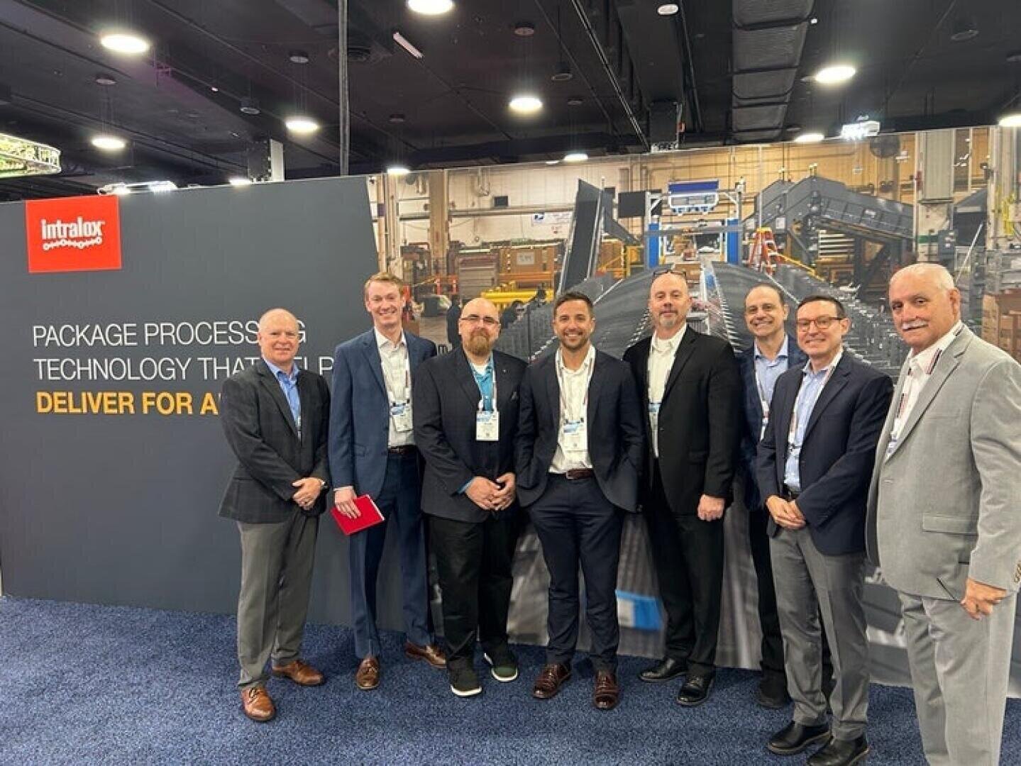 Acht Männer in Geschäftskleidung stehen lächelnd vor einem Messestand von Intralox mit einem großen Display über Verpackungsverarbeitungstechnologie und -ausrüstung in einem Kongresszentrum.