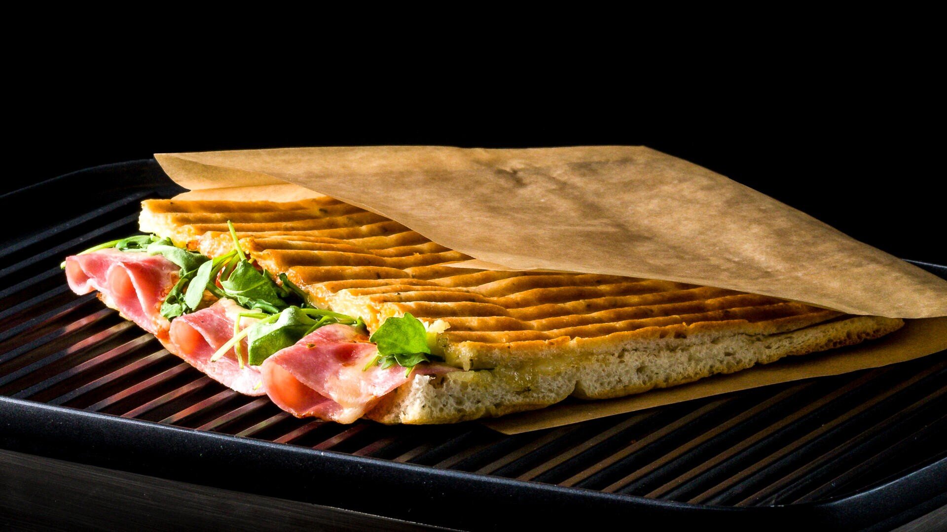 A toasted panini filled with ham and arugula is partially wrapped in brown parchment paper, resting on a ridged grill pan against a black background.