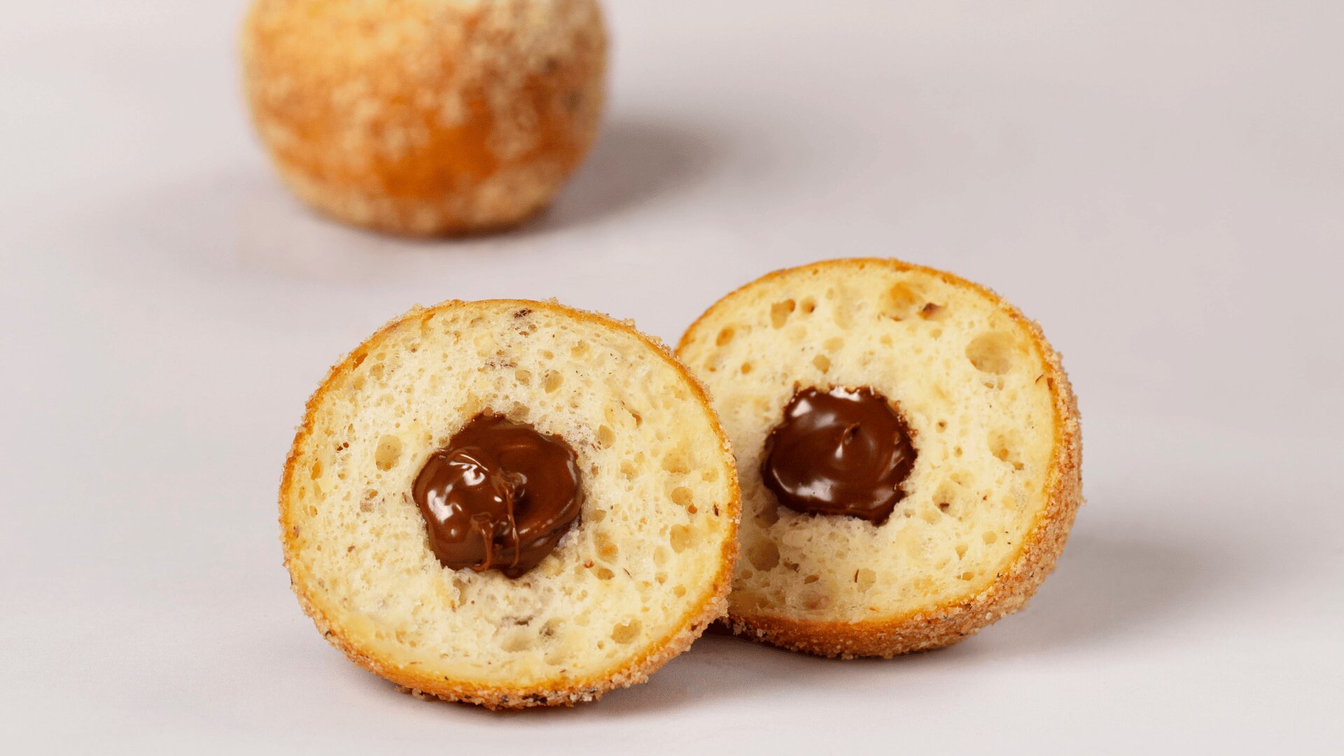 Ein rundes Gebäckstück ist in der Mitte durchgeschnitten, so dass im Inneren eine Schokoladencremefüllung zum Vorschein kommt. Ein weiteres ganzes Gebäckstück ist im Hintergrund zu sehen, alles auf einer hellen Fläche.