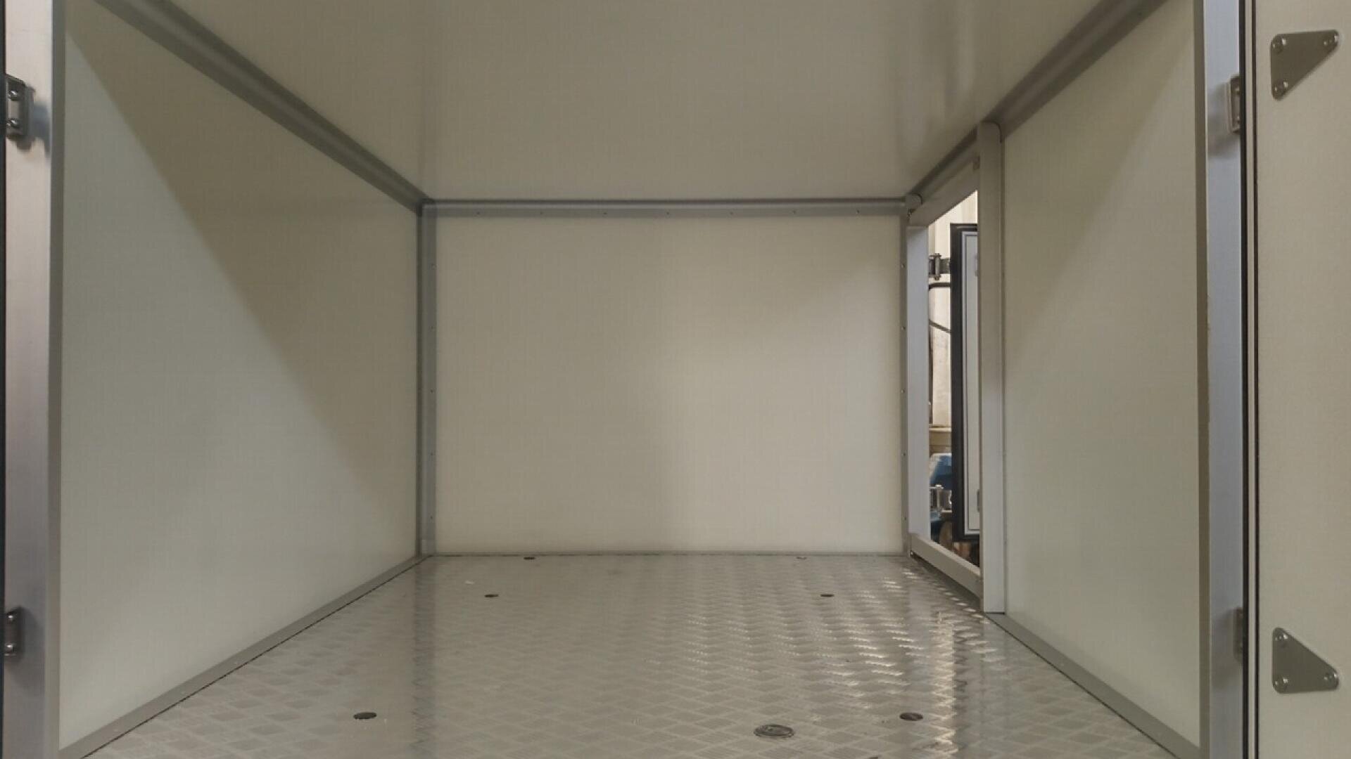 The image shows the interior of an empty, metallic, rectangular chamber with smooth white walls, a diamond-patterned floor, and an open door on the right side.