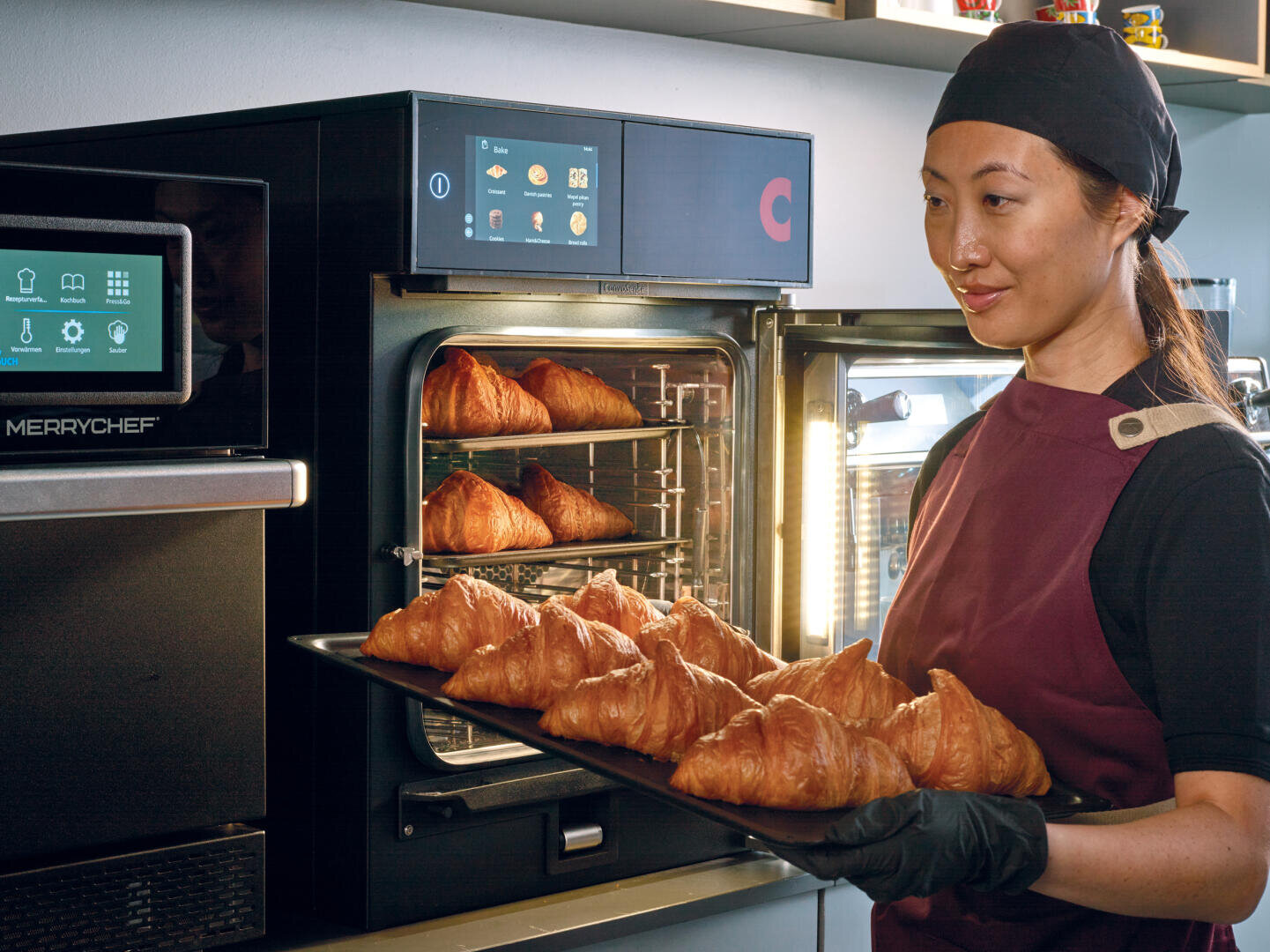 A woman wearing a chefs uniform and gloves holds a tray of fresh croissants in front of a commercial oven, with more croissants baking inside. She stands in a modern kitchen with digital cooking equipment.