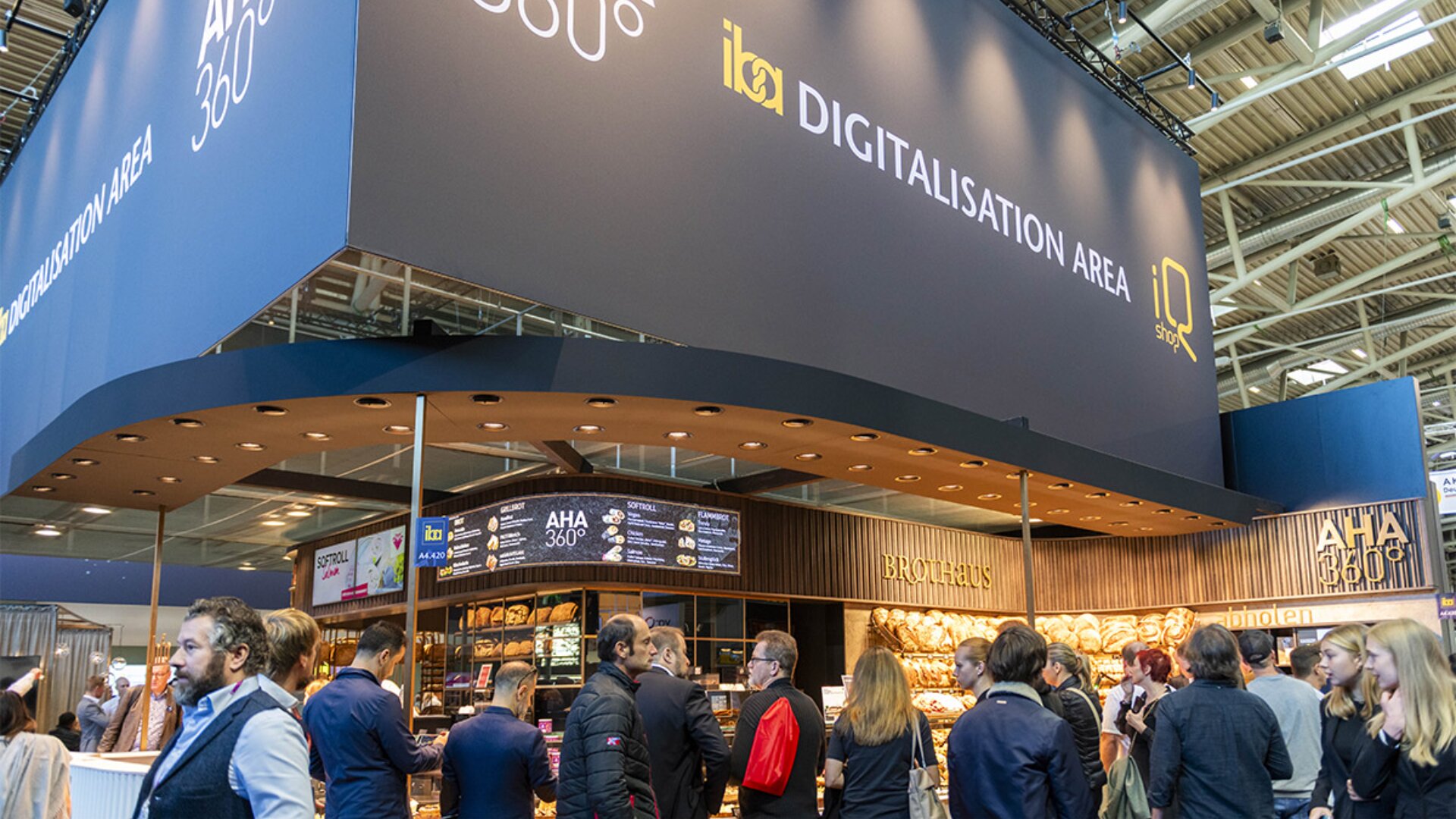 Eine belebte Ausstellungshalle mit Menschen, die um einen Bäckereistand unter einem großen Schild mit der Aufschrift DIGITALISIERUNGSBEREICH versammelt sind. Brotauslagen sind zu sehen, und der Veranstaltungsort wirkt modern und lebendig.