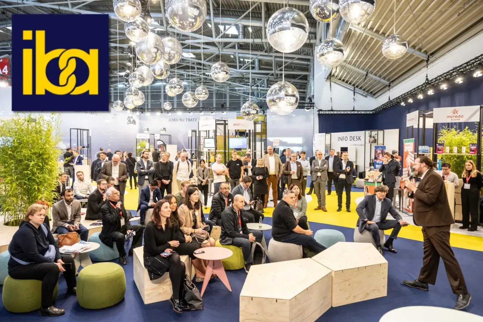 Ein Redner wendet sich an ein sitzendes und stehendes Publikum bei einer modernen Indoor-Veranstaltung oder -Konferenz, mit dekorativen hängenden Kugeln und einem großen "iba"-Logo in der oberen linken Ecke. Die Atmosphäre ist lebendig und professionell.