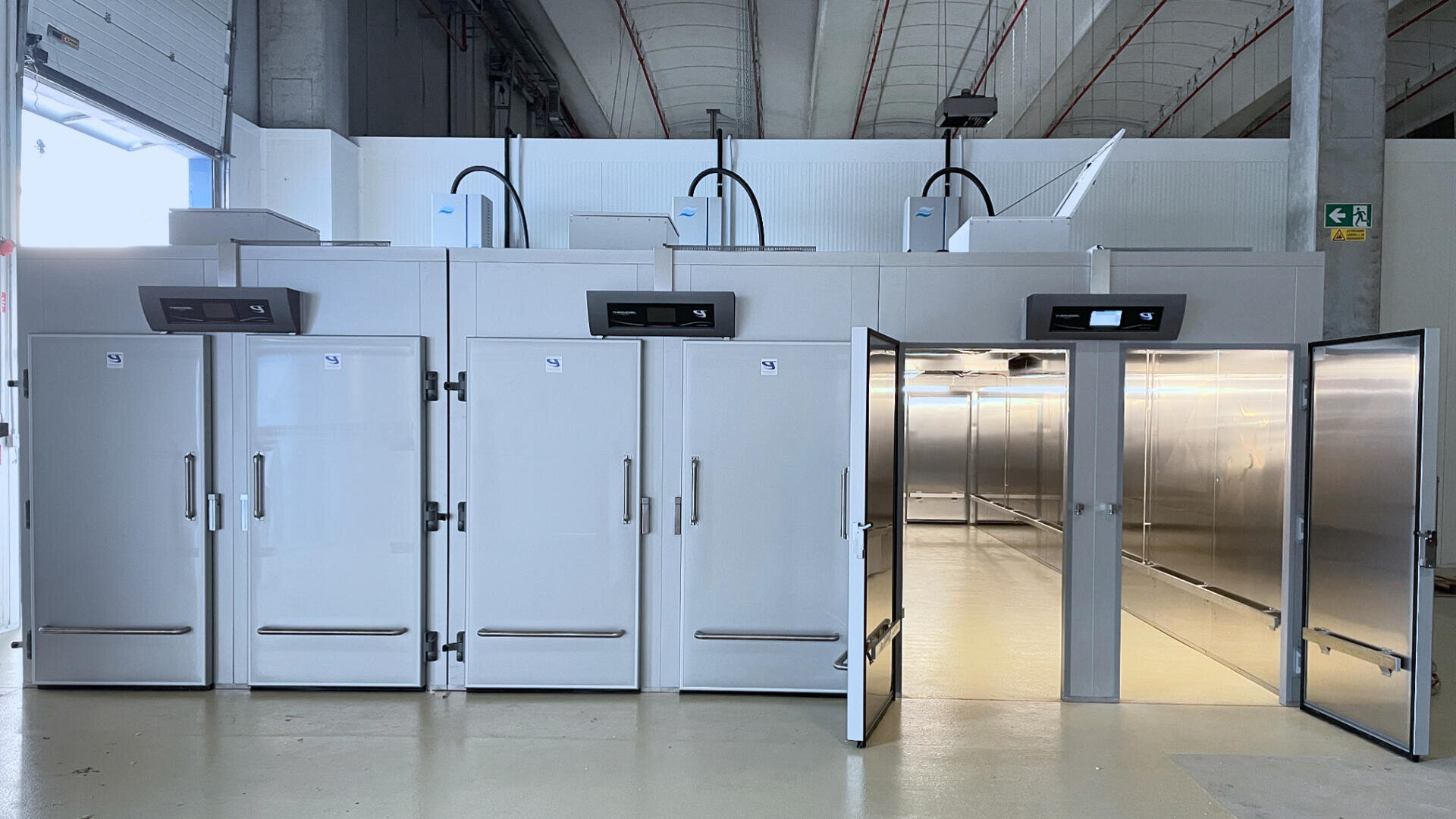 A row of large, industrial walk-in refrigerators or freezers with gray doors; one unit is open, revealing a spacious, empty interior with metallic walls and ceiling-mounted cooling units.