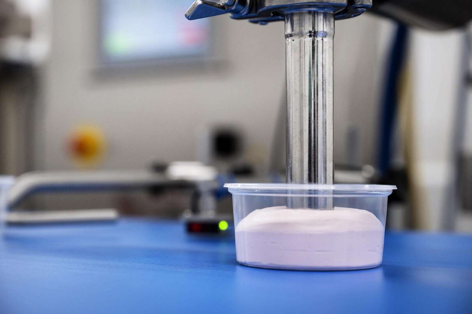 A metal dispenser fills a clear plastic container with a light pink, creamy substance on a blue surface in an industrial setting.