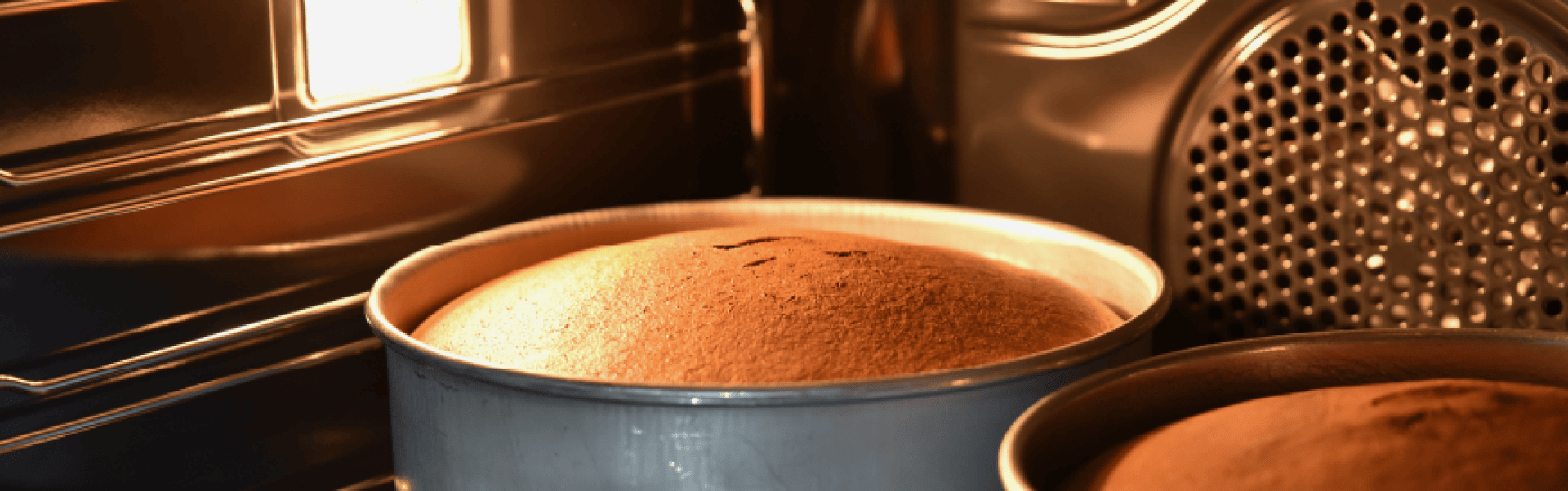 Two round cake pans with rising cakes inside an oven, illuminated by an oven light. The cakes are golden brown and appear to be nearly finished baking.