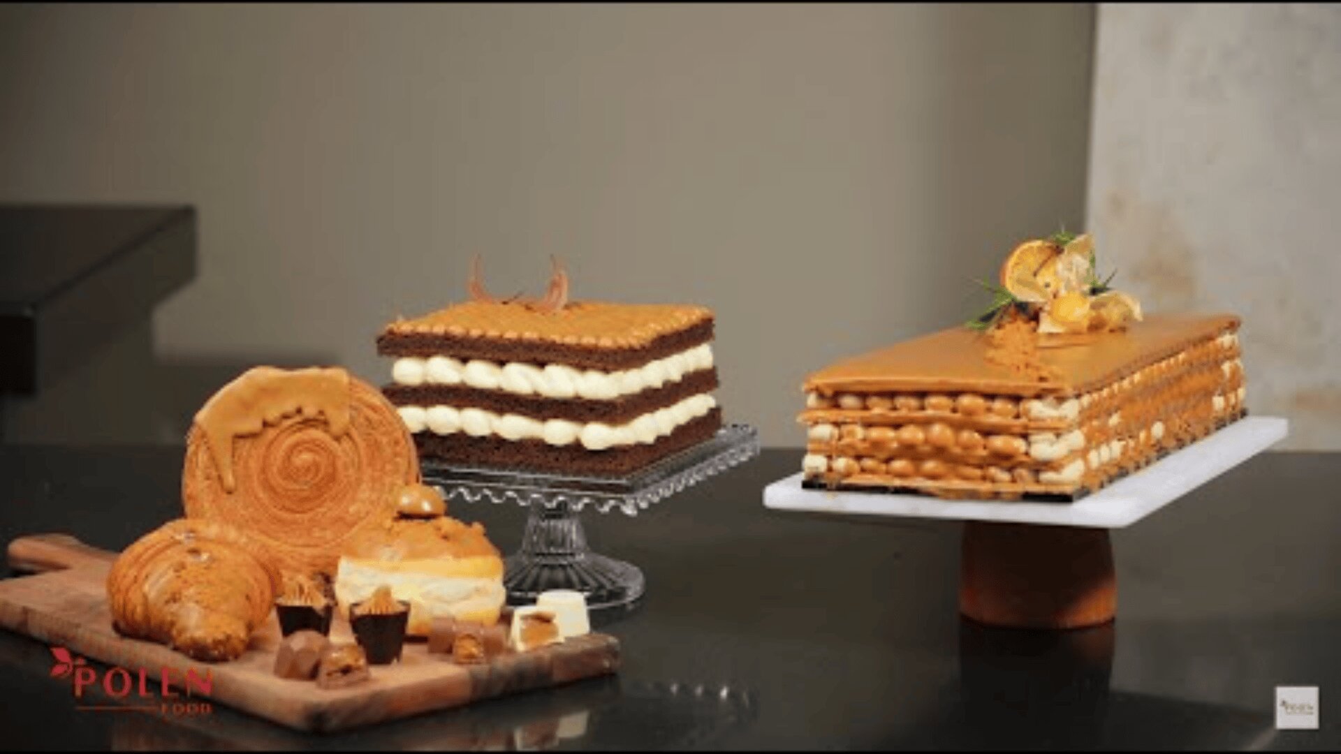 Auf einer dunklen Theke werden drei elegante Tortenschichten und eine Auswahl an Gebäck präsentiert. Die Torten sind mit Sahne und Karamell überzogen, während das Gebäck aus Croissants und gewirbelten Brötchen auf einem Holzbrett besteht.