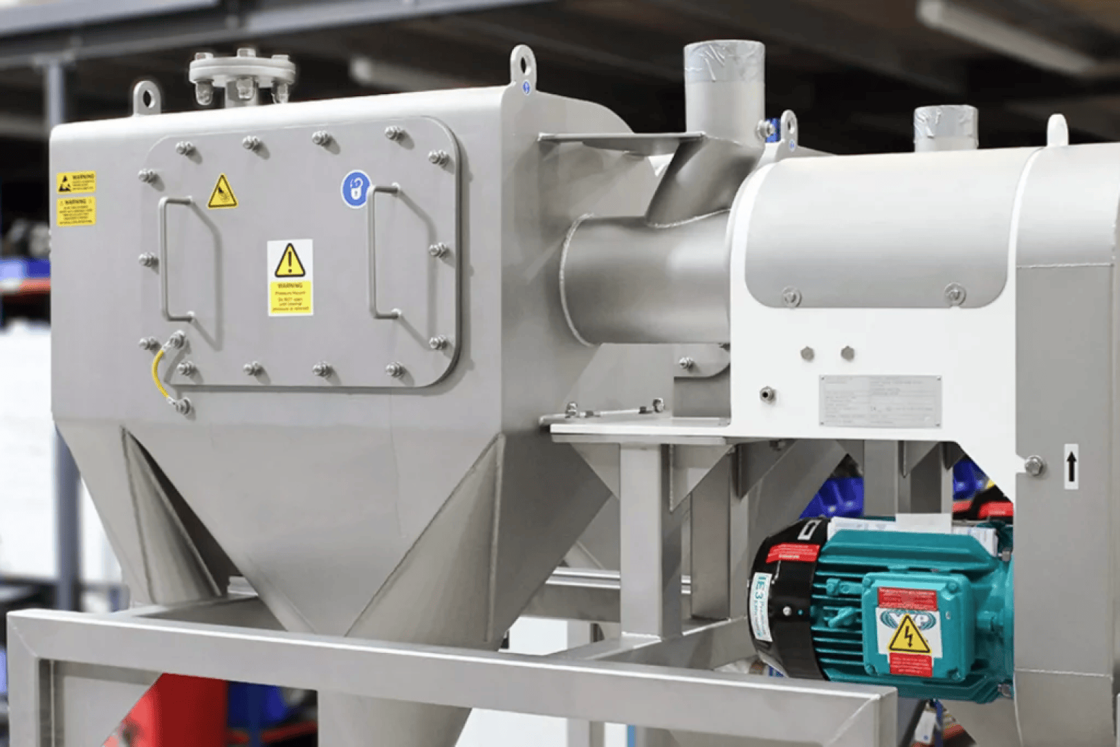 Industrial machines made of stainless steel with warning signs, mounted on a frame. A green electric motor is attached to one side. The equipment appears to be used for processing or manufacturing in an industrial environment.