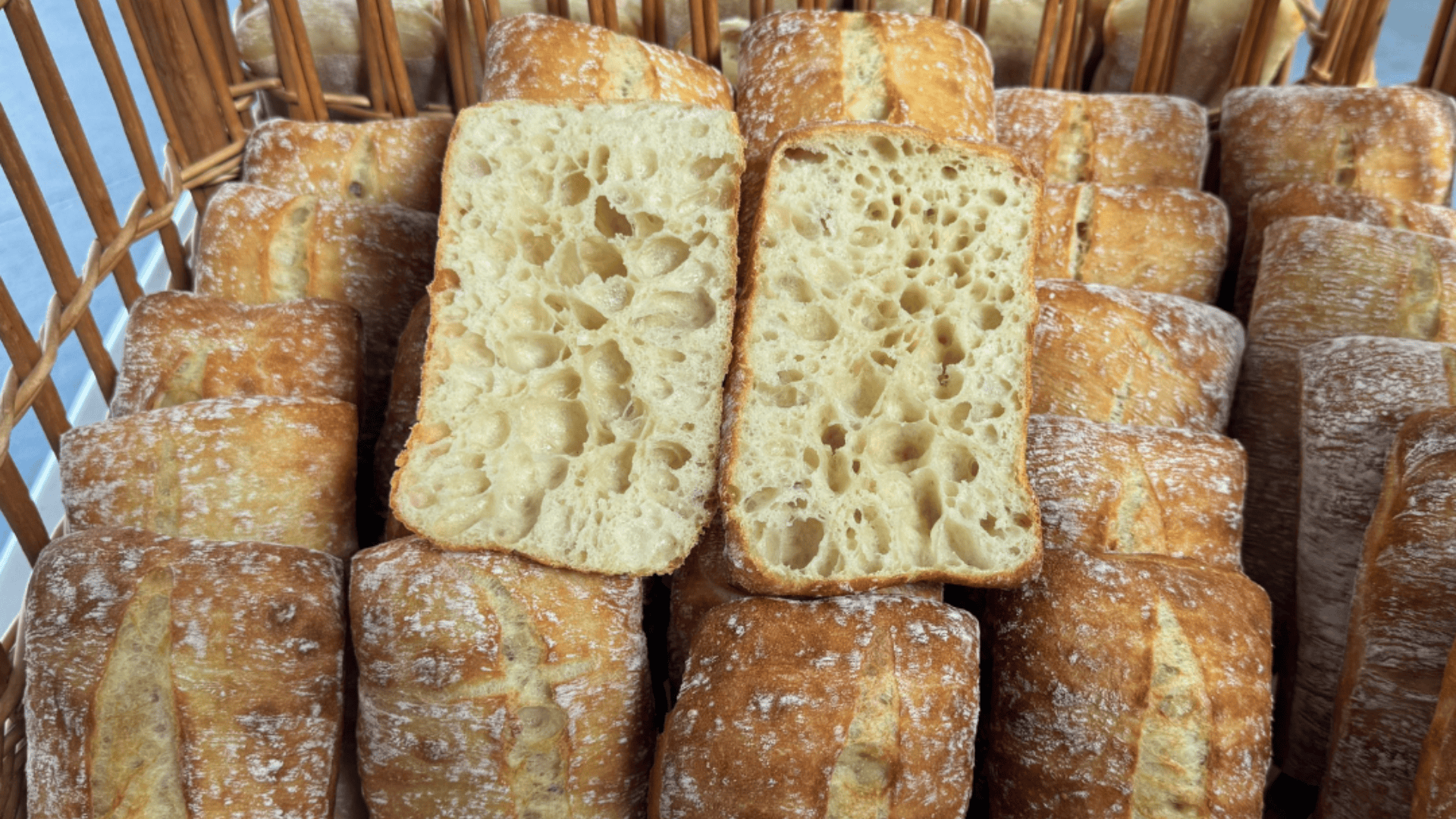 Rechteckige Ciabatta-Brötchen werden in einem Korb gestapelt. Zwei Brötchen werden aufgeschnitten, so dass die luftige, mit Löchern versehene Krume zum Vorschein kommt, während die goldbraune Kruste leicht mit Mehl bestäubt ist.
