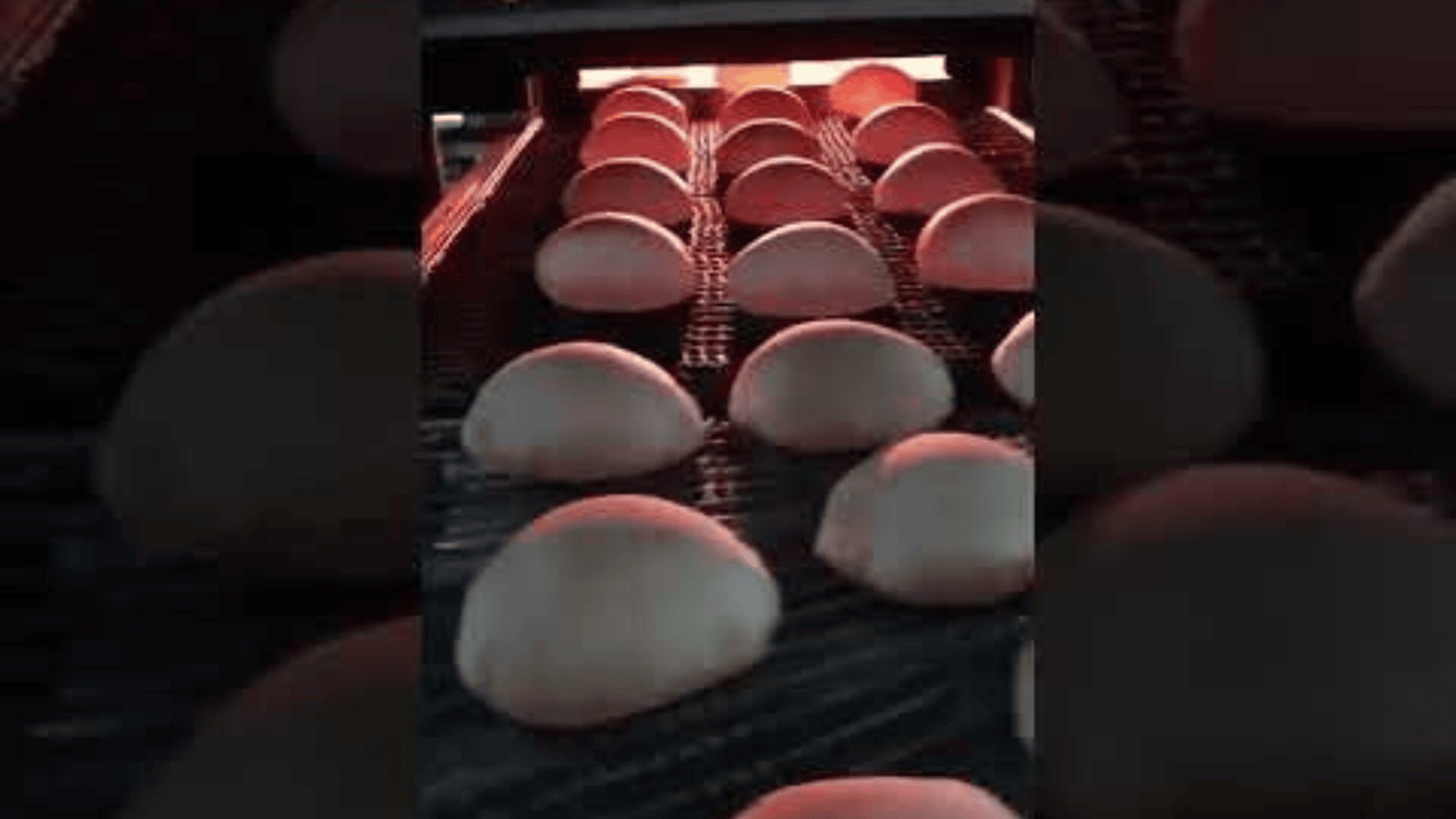 Rows of round bread dough rise on a conveyor belt and move towards an industrial oven, which glows warmly in the background.