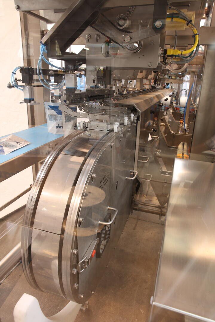 A close-up view of industrial packaging machinery with metallic parts, blue cables, and conveyor components inside a transparent protective enclosure.