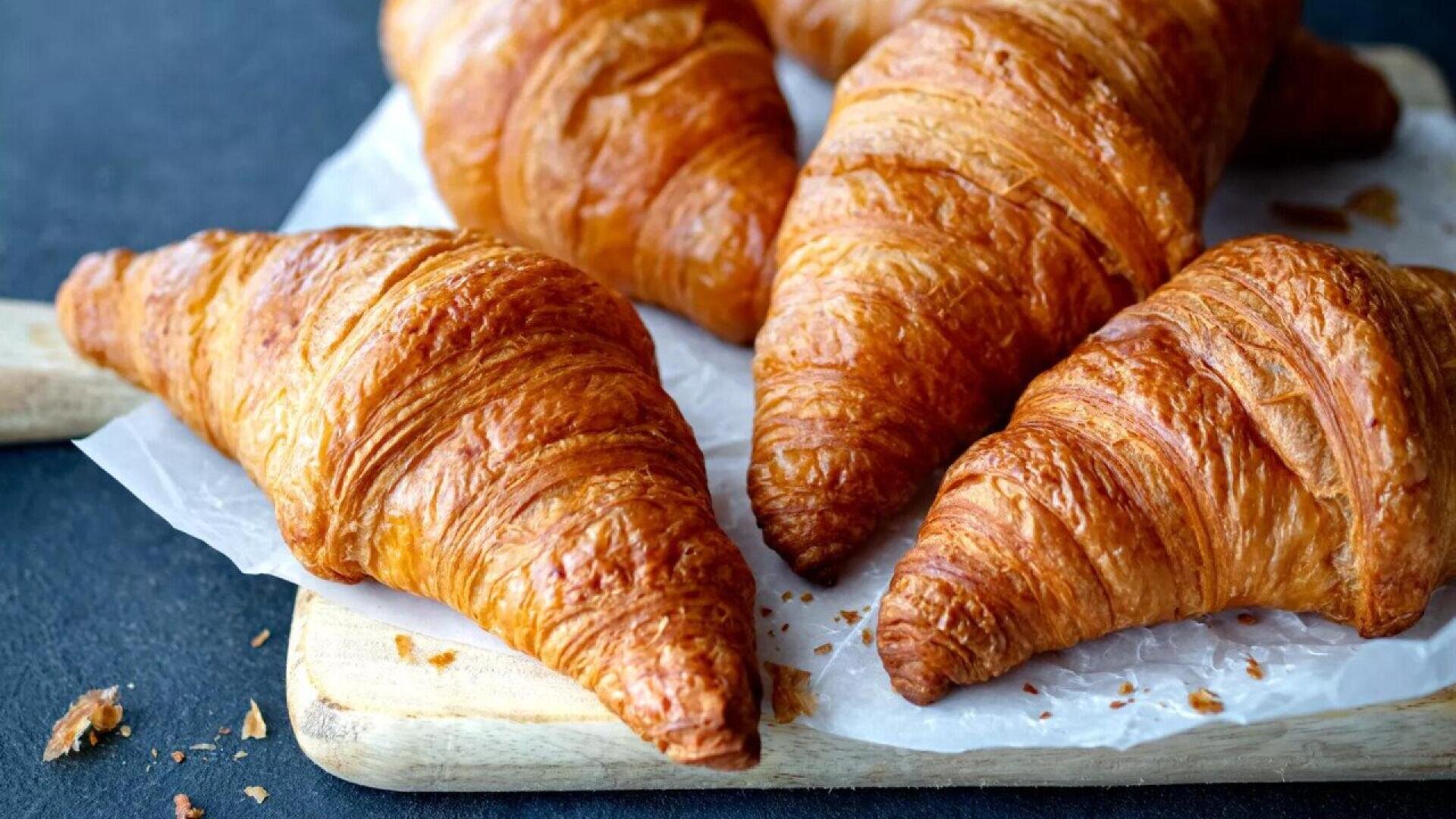 Four golden, flaky croissants are arranged on a piece of parchment paper atop a wooden board, with a dark background and a few crumbs scattered nearby.