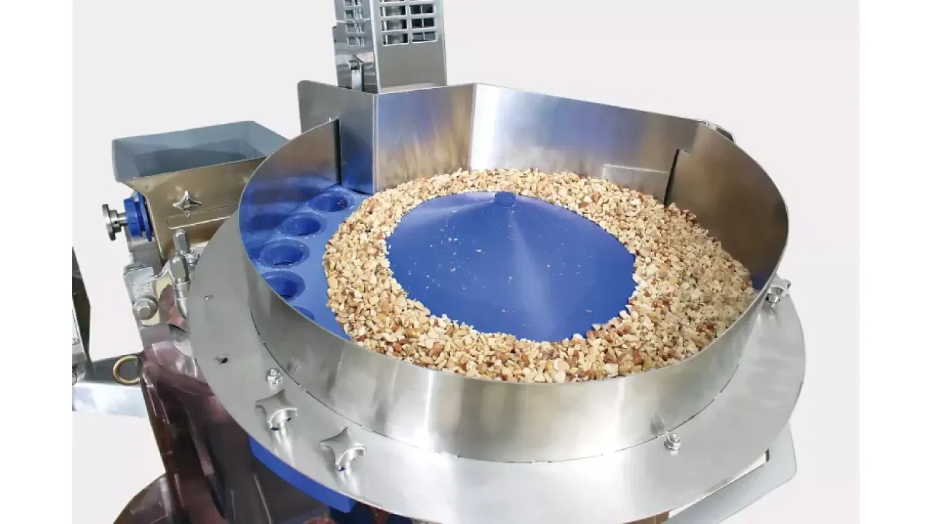 Eine Industriemaschine aus rostfreiem Stahl mit einem kreisförmigen blauen Förderband, das kleine Stücke von gehackten Nüssen oder Samen in einem Ring um die Mitte sortiert oder verarbeitet.
