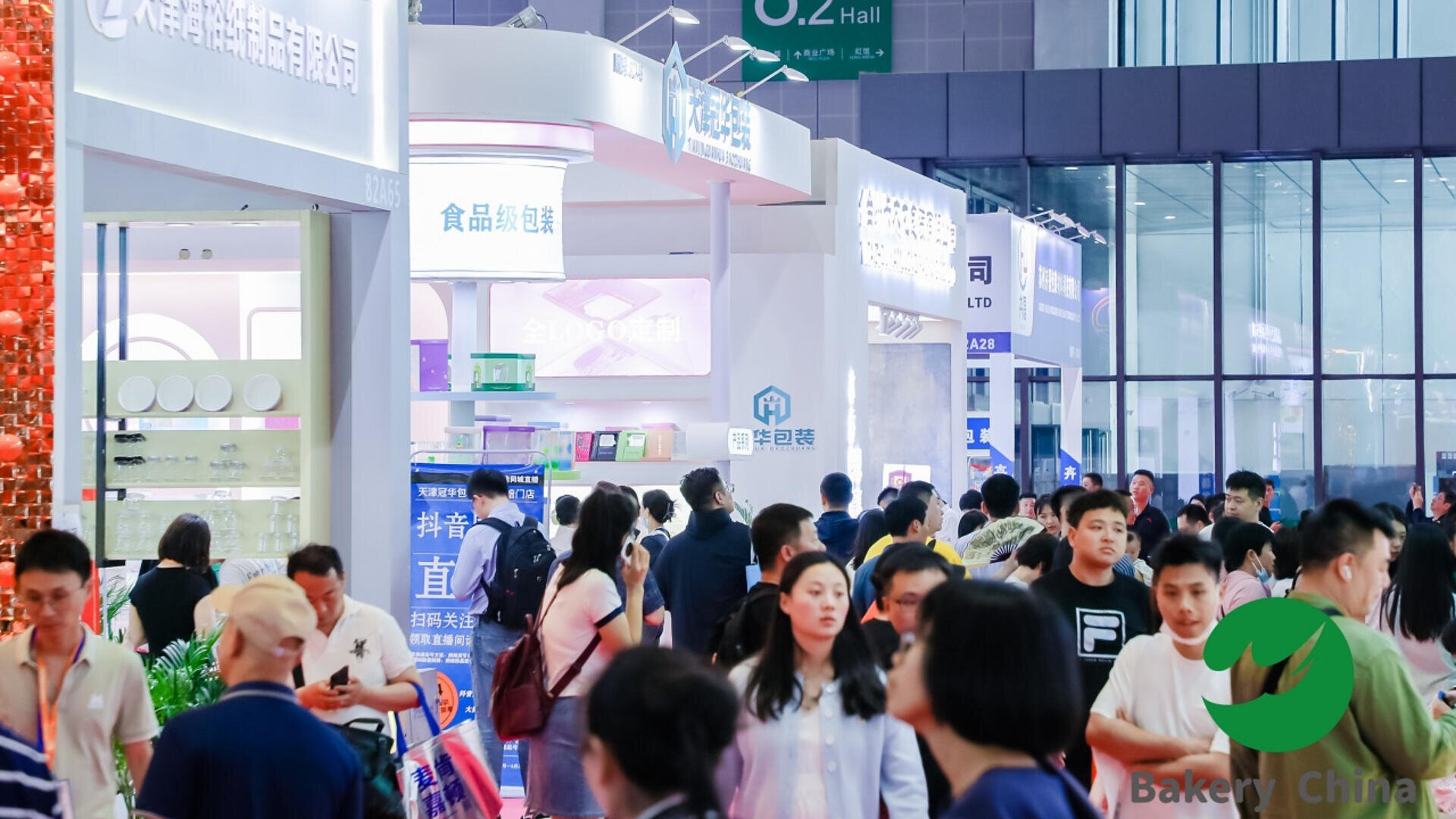 Large crowd of people walk through a busy, brightly lit exhibition hall at a trade show. Booths with signs in Chinese and displays line both sides. The Bakery China logo appears in the lower right corner.