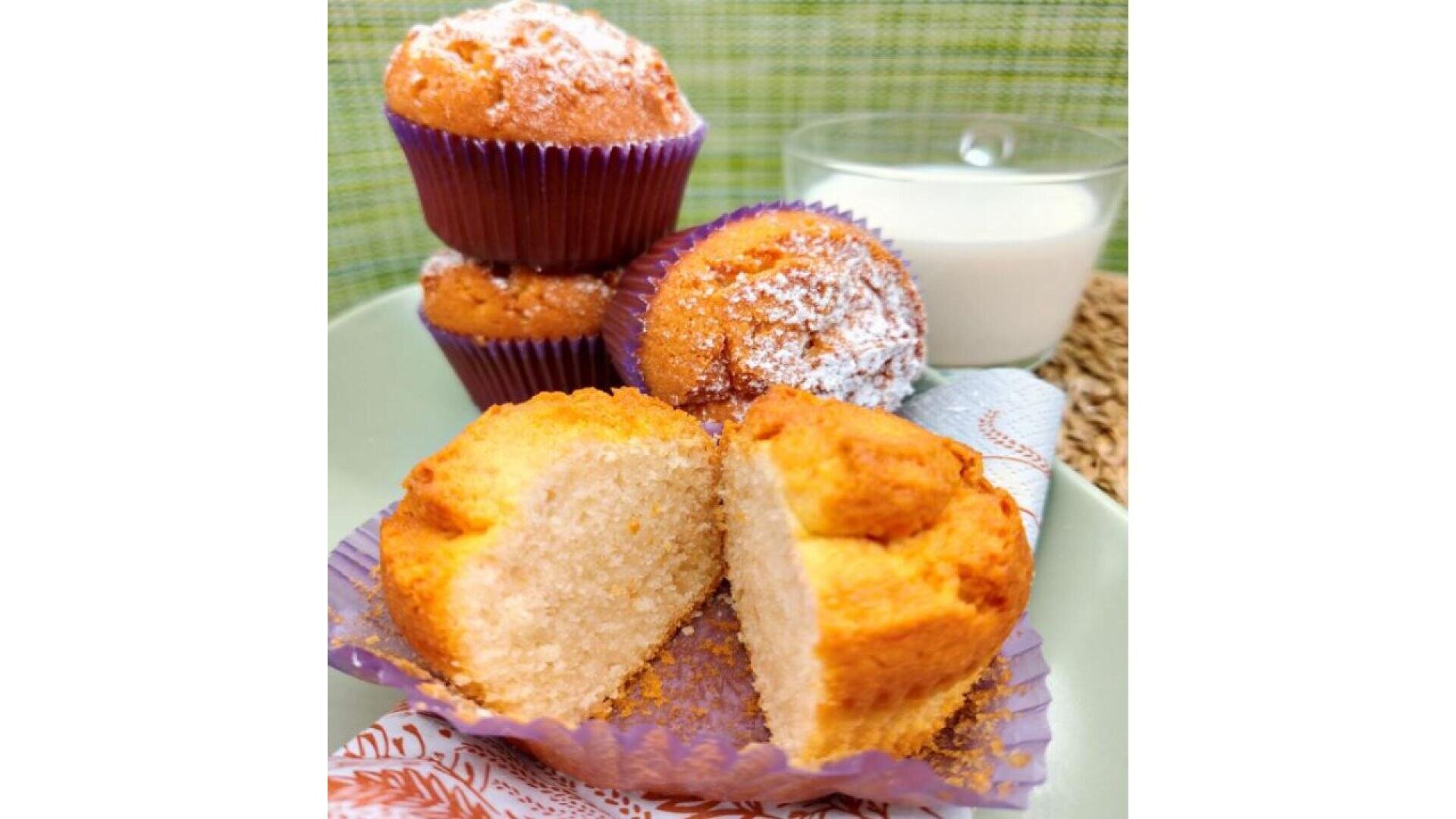 A plate with cut and whole golden muffins in purple wrappers, some dusted with powdered sugar, next to a glass of milk on a green-patterned background.