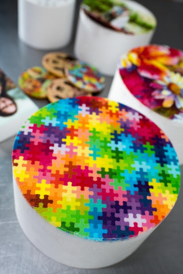 Ein runder Kuchen ist mit einem essbaren Bild aus bunten, ineinandergreifenden Puzzleteilen verziert. Weitere dekorierte Kuchen und Kekse sind im unscharfen Hintergrund auf einer metallischen Oberfläche zu sehen.