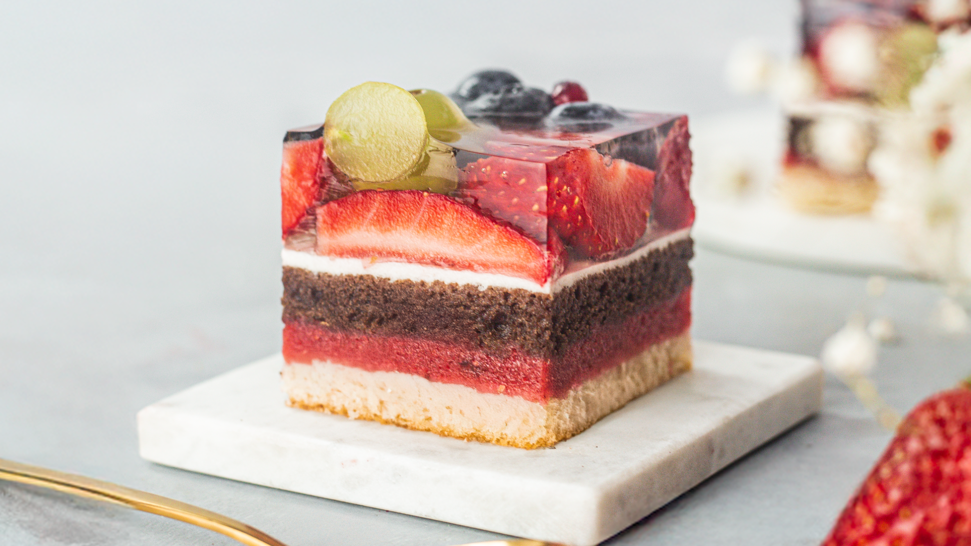 A square slice of layered cake with fruit topping, including grapes and berries. The cake has layers of sponge, chocolate, cream, and jelly, and is served on a small marble plate.