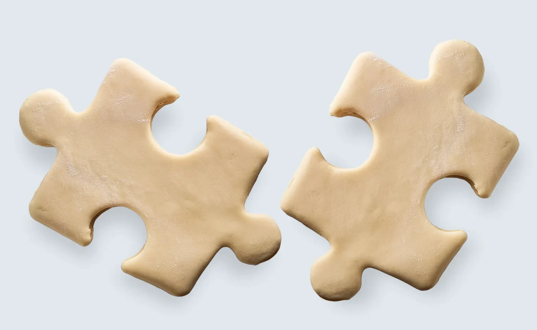 Zwei rohe, puzzleteilförmige Kekse aus hellem Teig liegen nebeneinander auf einem hellen Untergrund.