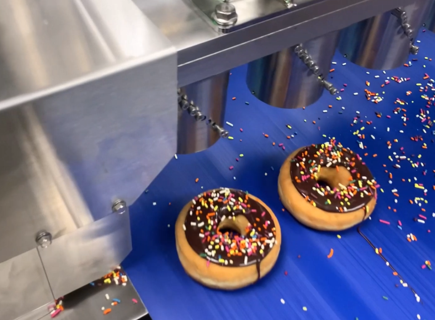 Two chocolate-glazed donuts with colorful sprinkles move along a blue conveyor belt under a metal machine in a factory setting.