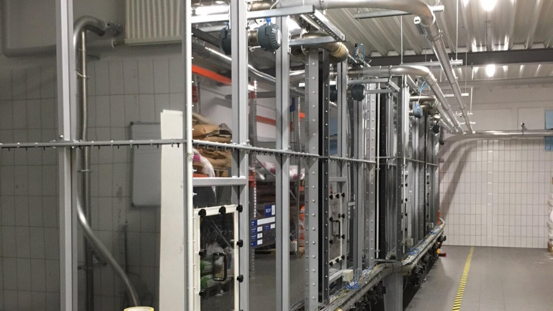 A large industrial machine with metal pipes and framework in a tiled room, possibly part of a factory or processing plant, with overhead lighting and yellow safety markings on the floor.