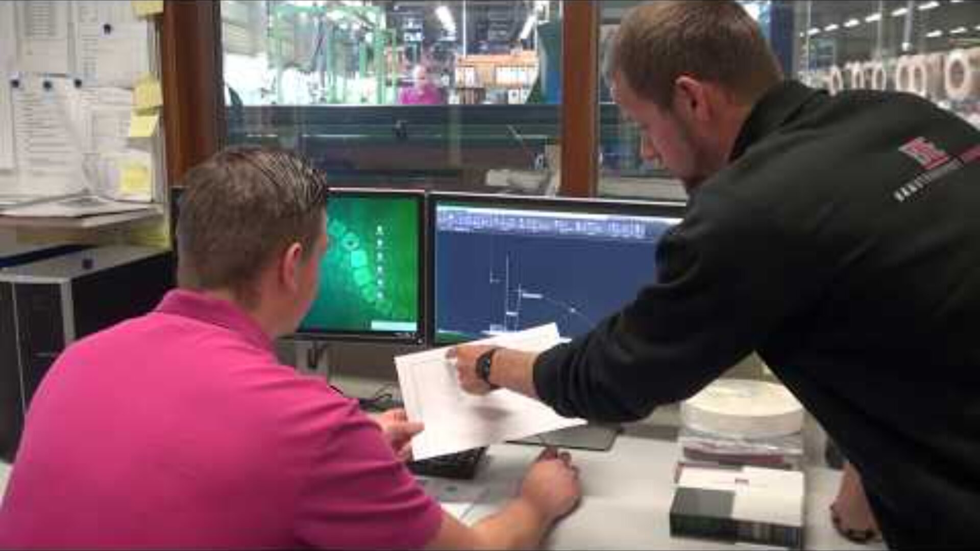 Two men work at a desk with two monitors displaying technical diagrams. One man sits and looks at the screens while the other stands, pointing at a document in his hand, discussing the information.