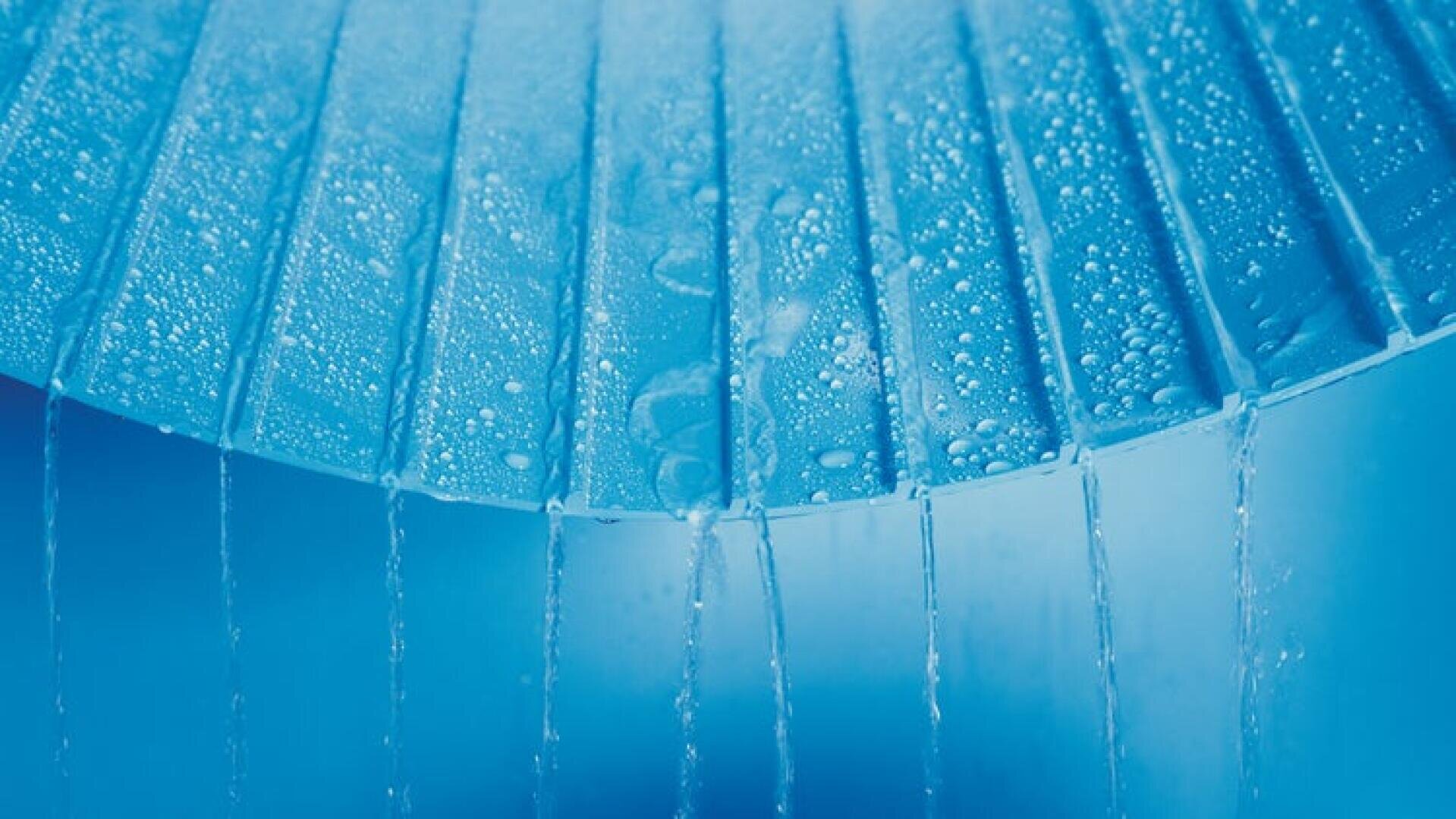 Close-up of water droplets on tilted blue slats with thin streams of water flowing down between the gaps against a soft blue background.