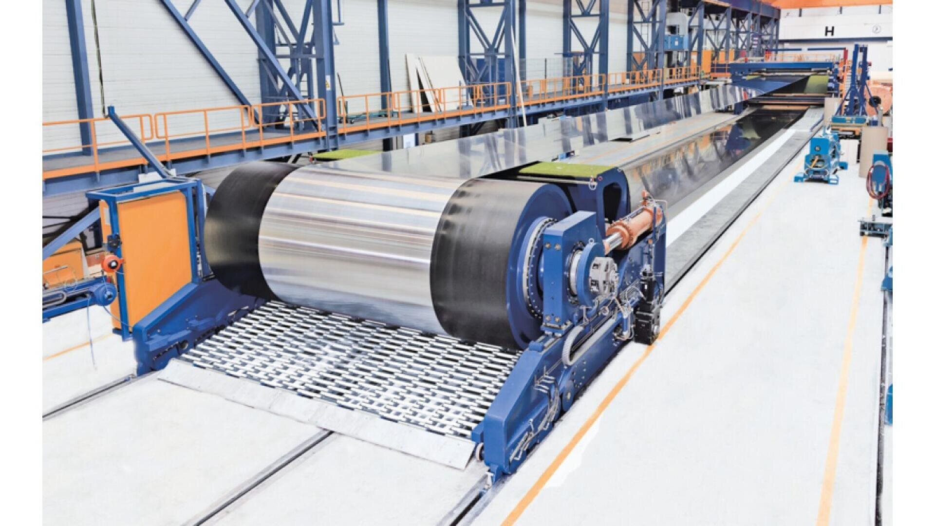 A large industrial machine in a factory setting is unwinding and processing a wide sheet of shiny metallic material along a conveyor system. Blue support structures and safety railings are visible around the equipment.