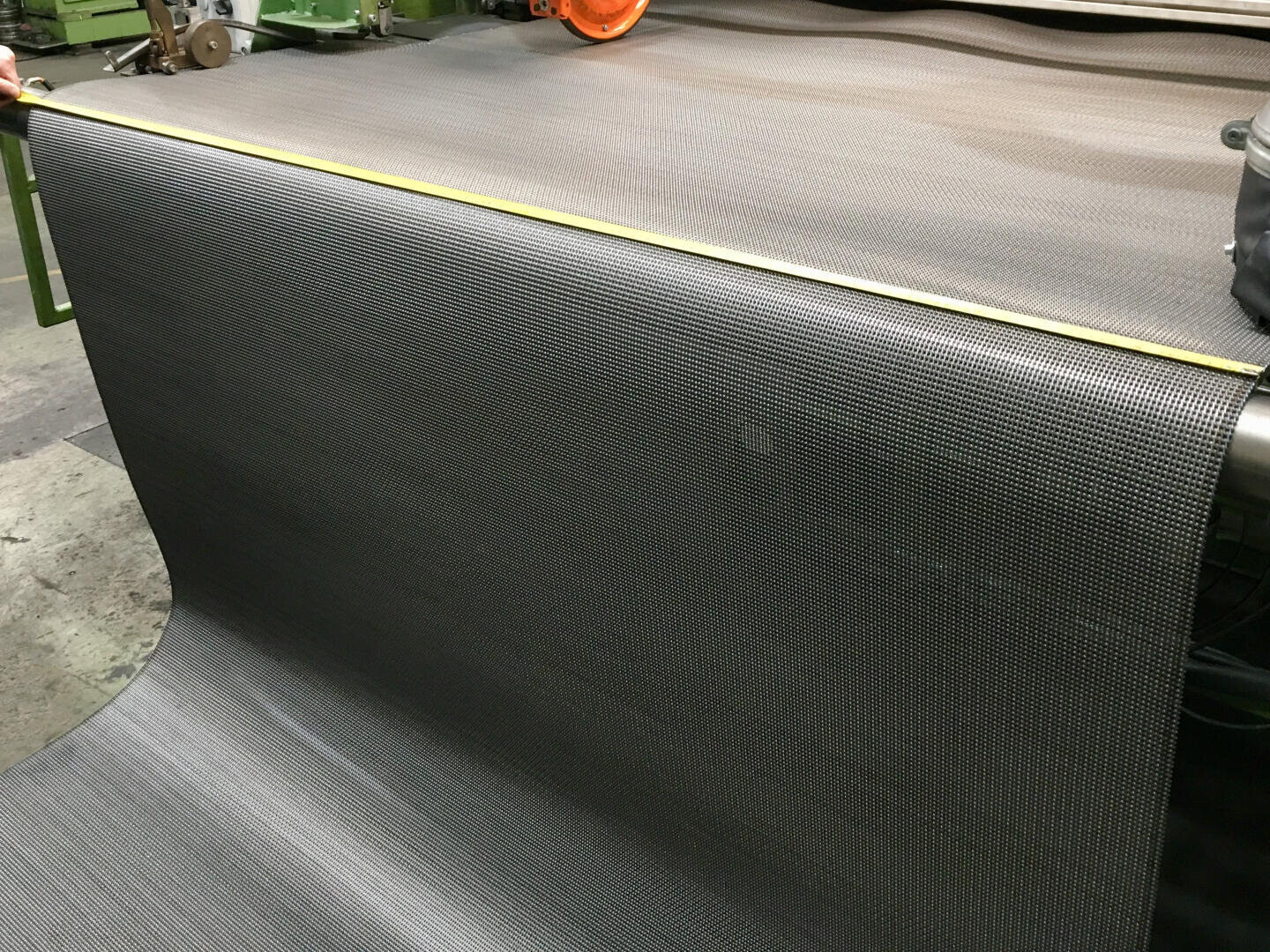 Ein großes Stück schwarzes Industriegewebe wird mit einem gelben Maßband auf dem Boden einer Fabrik gemessen, im Hintergrund sind Maschinen zu sehen.