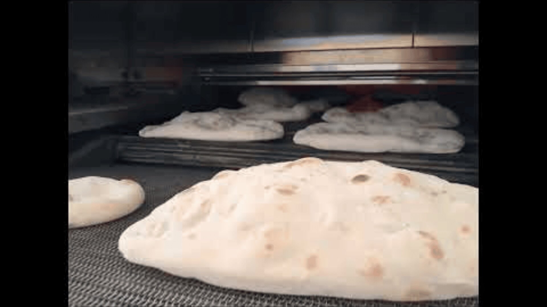 Several pieces of flatbread are baking and puffing up inside a commercial oven, with one piece prominently in the foreground.