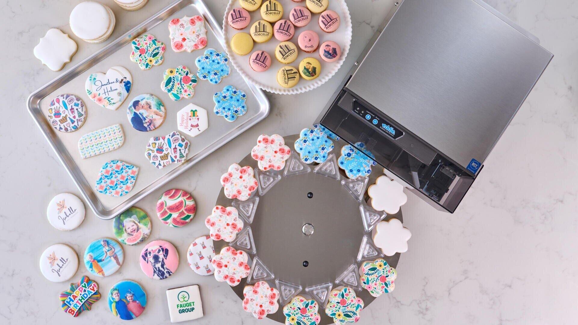 Various colorful cookies, some with printed photos and motifs, are arranged on trays and around a round tray. A metal printing machine stands next to the cookies on a marble slab.