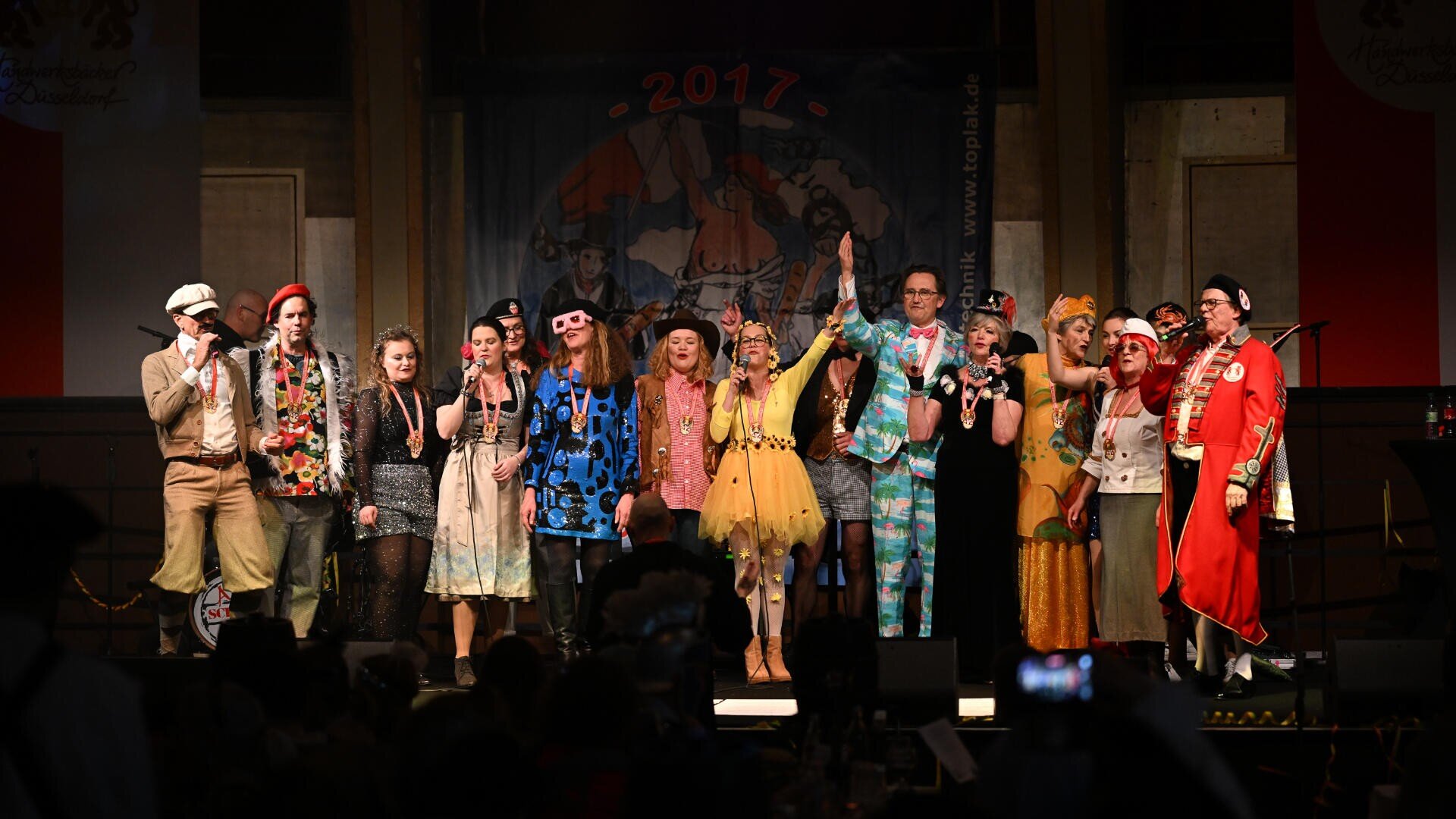 Eine Gruppe von Menschen in bunten, vielseitigen Kostümen steht gemeinsam auf der Bühne, einige winken oder halten Mikrofone in der Hand, unter einem Banner mit der Aufschrift 2017, während einer festlichen Veranstaltung mit einem Publikum im Vordergrund.