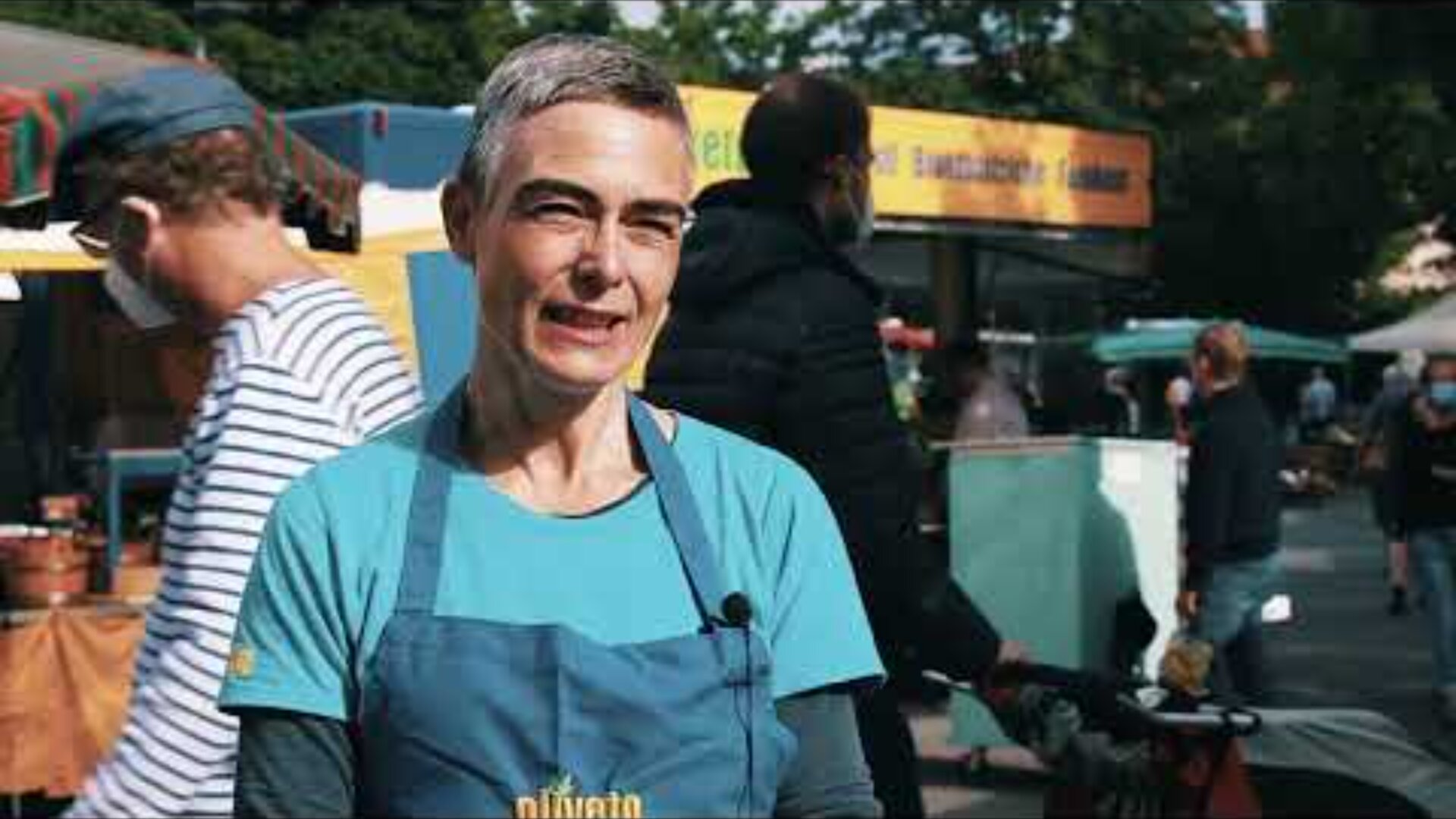 Eine Person mit kurzen grauen Haaren, die ein blaues Hemd und eine Schürze trägt, steht im Freien auf einem belebten Markt mit Menschen und bunten Verkaufsständen im Hintergrund.