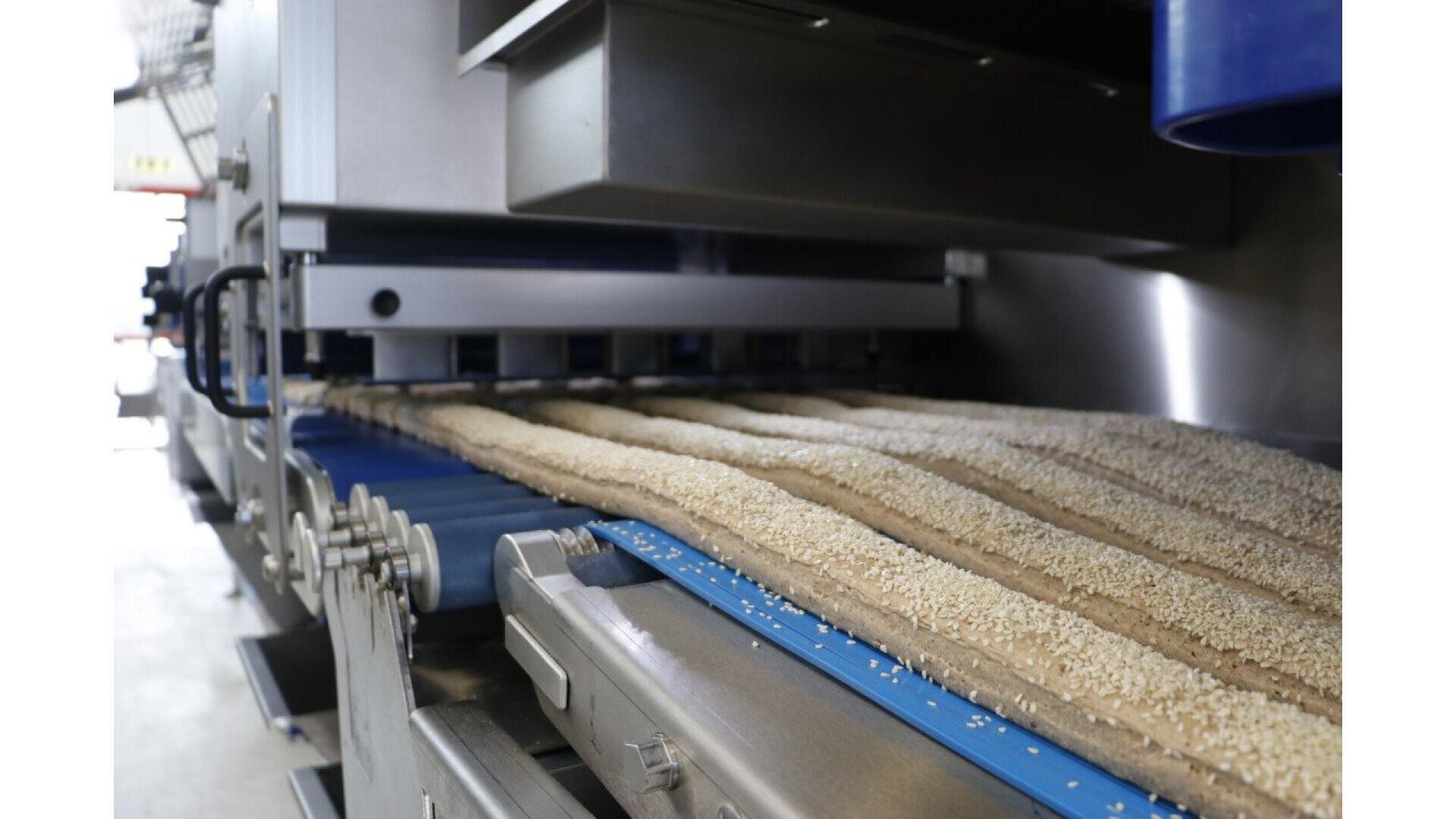 A close-up view of an industrial machine processing dough strips coated with sesame seeds on a conveyor belt in a food production facility.