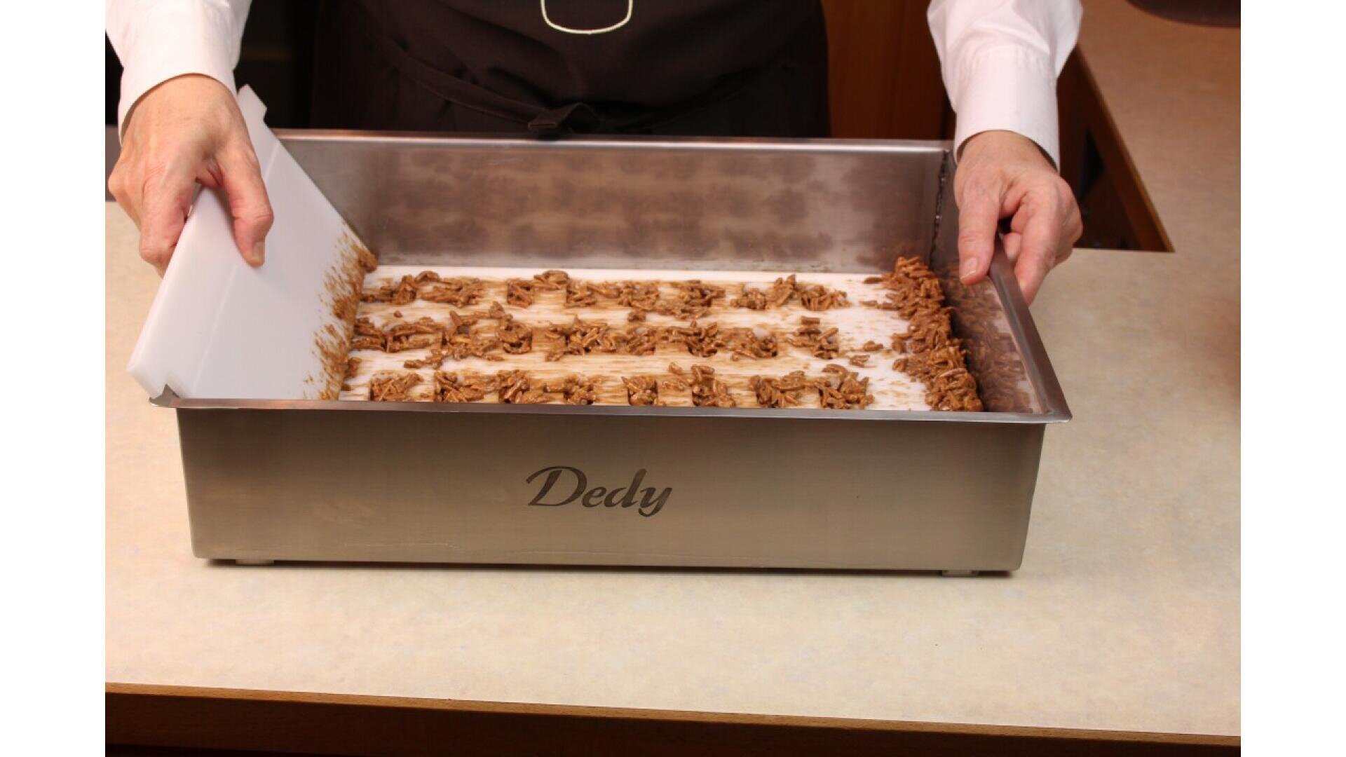 A person uses a white scraper to shape rows of chocolate in a rectangular metal tray labeled Dedy on a light countertop.