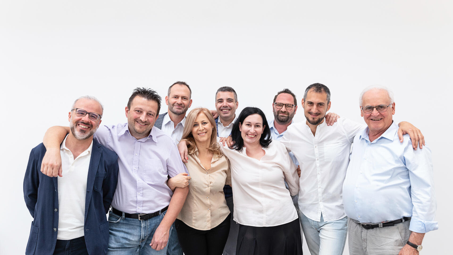 Eine Gruppe von acht Erwachsenen, fünf Männern und drei Frauen, steht Seite an Seite vor einem weißen Hintergrund. Sie lächeln und wirken freundlich, manche verschränken die Arme, andere legen sich gegenseitig die Hände auf die Schultern.